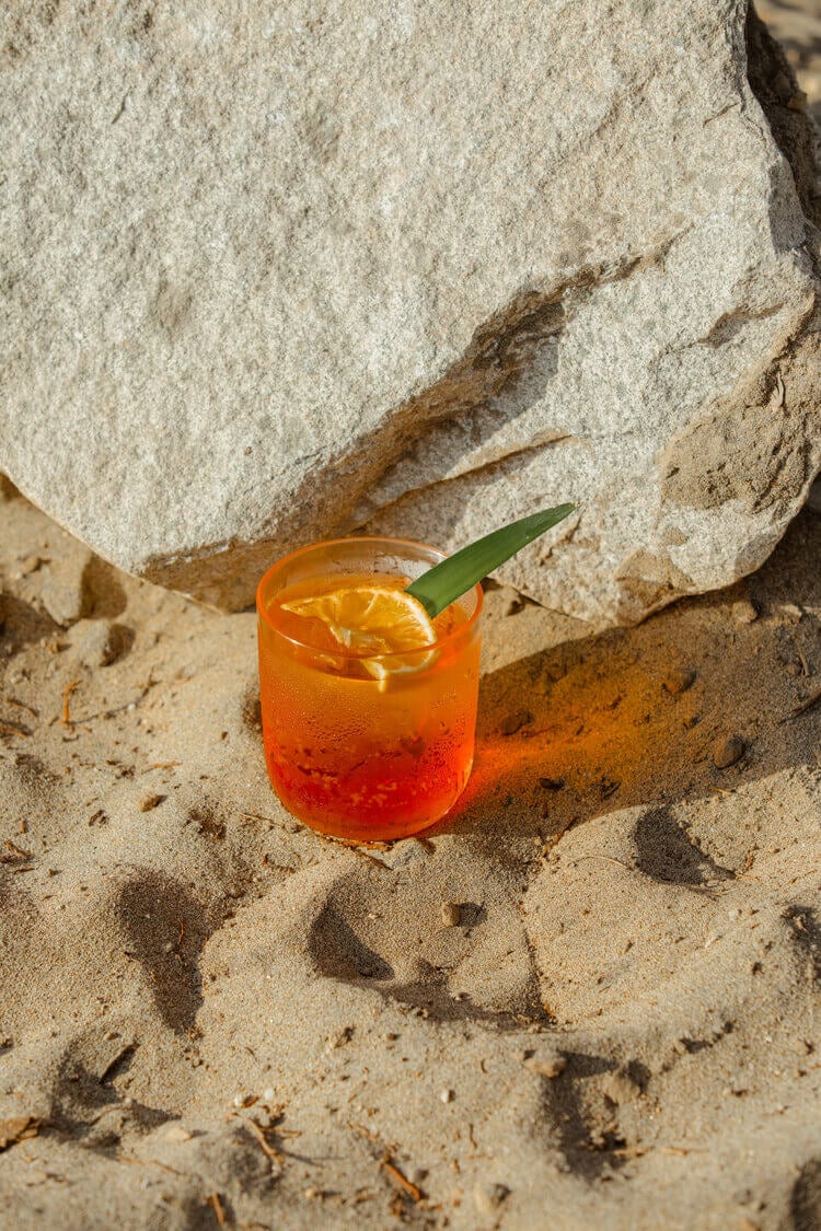 Club de Playa, Mexico — coral cocktail with citrus slice and palm leaf garnish on sand beside weathered stone, tropical beach setting with dappled sunlight