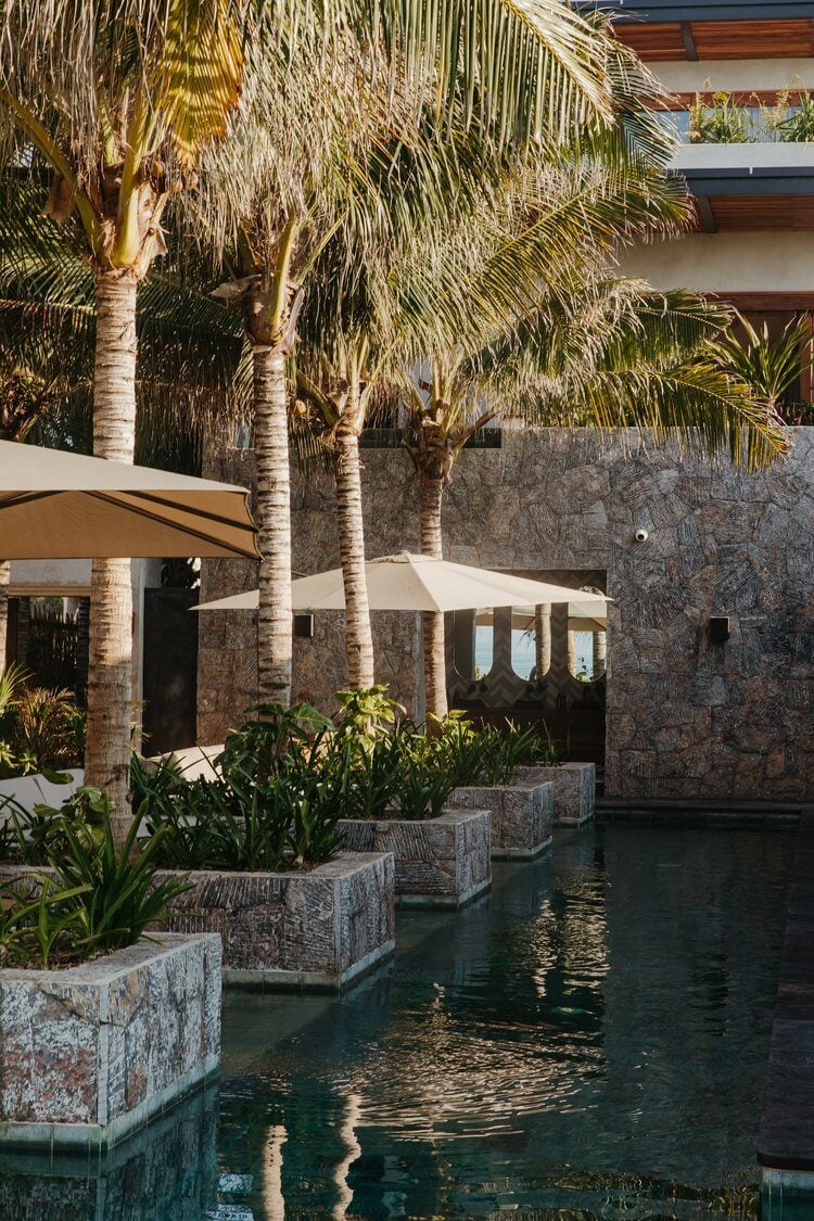 Hotelito MUSA, Mexico — Stone courtyard with palm trees, reflected water channel, canvas shade structures, and modernist architecture in warm sunlight.