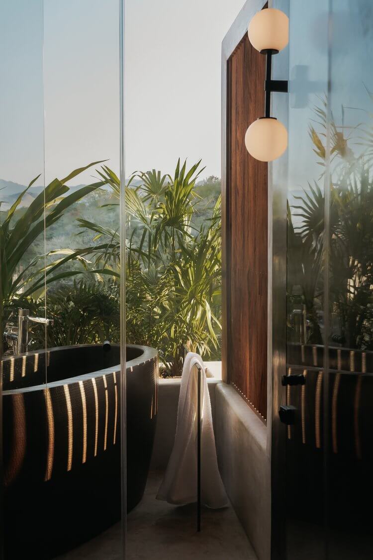 Hotelito MUSA, Mexico — Spa bathroom with curved black soaking tub, wooden wall paneling, glass partitions, tropical plants, and spherical pendant lighting overlooking lush landscape.