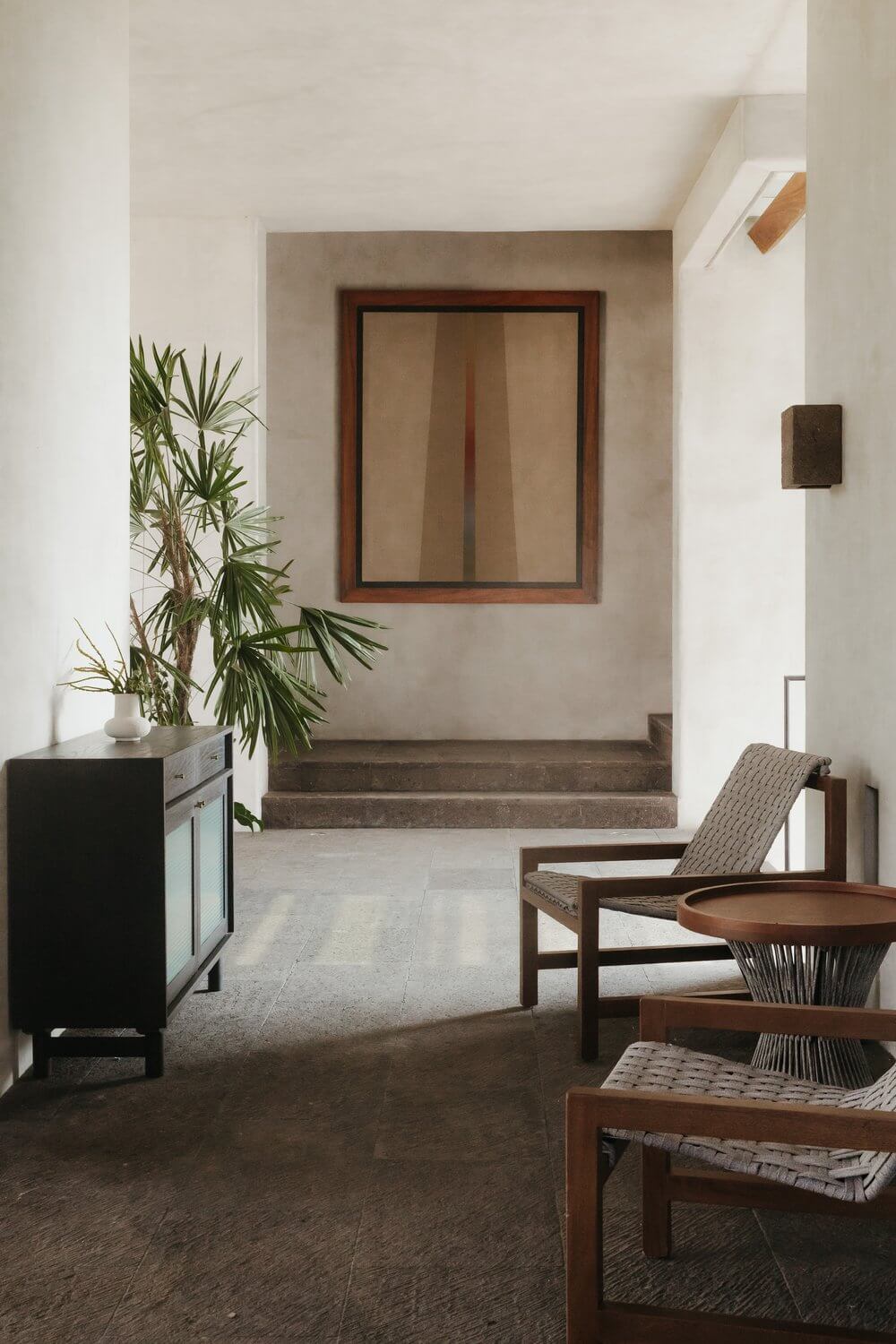 Hotelito MUSA, Mexico — Minimalist lobby with concrete floors, whitewashed walls, wooden furniture, potted palm, and framed mirror overlooking stepped entrance.