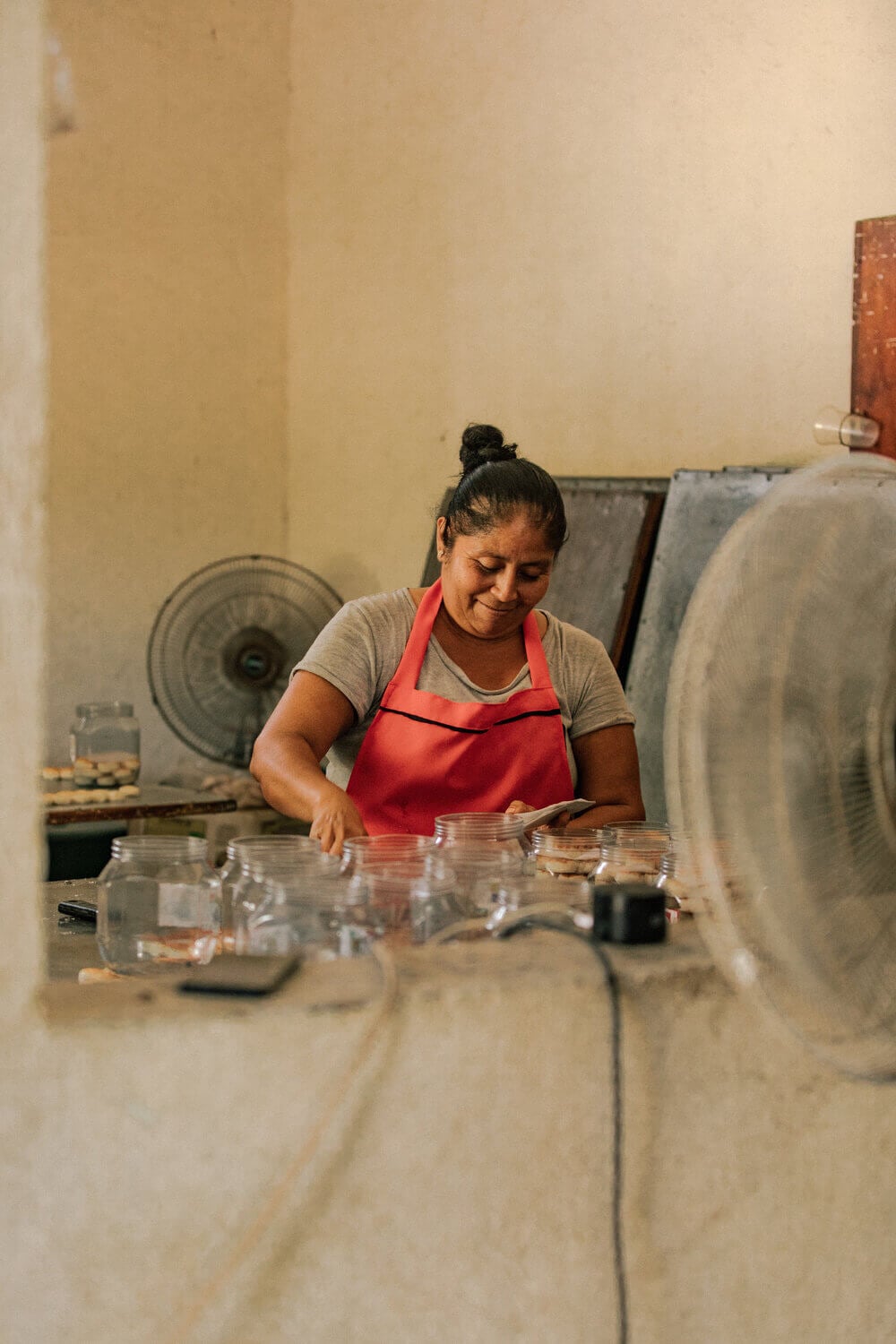 Hotelito MUSA, Mexico — Woman in red apron arranging glass jars in minimalist workshop with concrete surfaces and natural light.