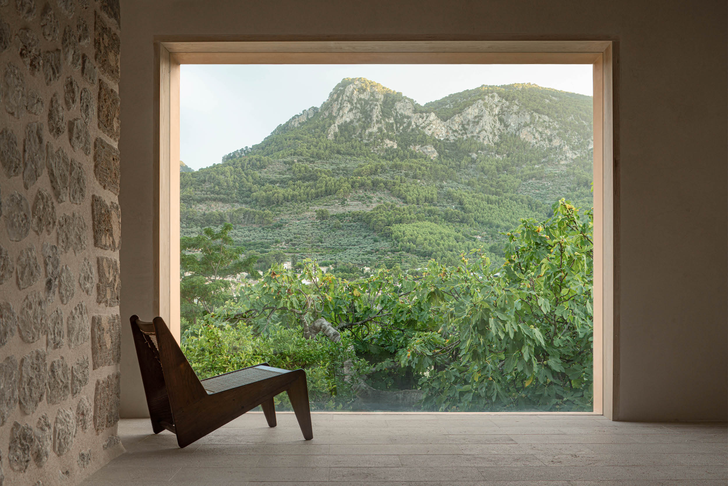 Can Gaspar, Balearic Islands — Dark wooden lounge chair facing expansive window overlooking terraced gardens, rocky peaks, and Mediterranean hillside landscape.