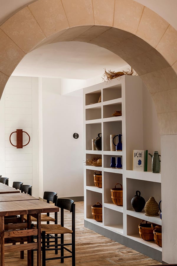 Menorca Experimental, Balearic Islands, Spain — Arched stone doorway opening to minimalist dining area with built-in shelving, natural light, terracotta tile floors, and curated ceramic vessels.