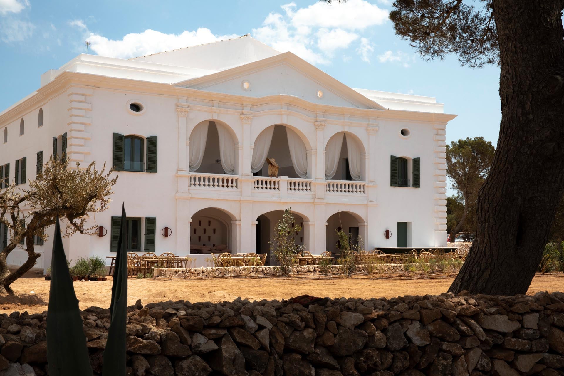 Menorca Experimental, Balearic Islands — Whitewashed neoclassical facade with arched loggias, stone balustrade, and terracotta shutters overlooking sandy courtyard with olive trees.