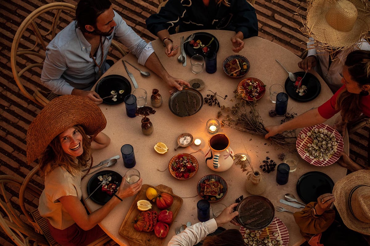 Menorca Experimental, Balearic Islands, Spain — Overhead view of four diners sharing rustic table with grilled vegetables, dark plates, dried botanicals, linen napkins, and candlelight on striped rug.