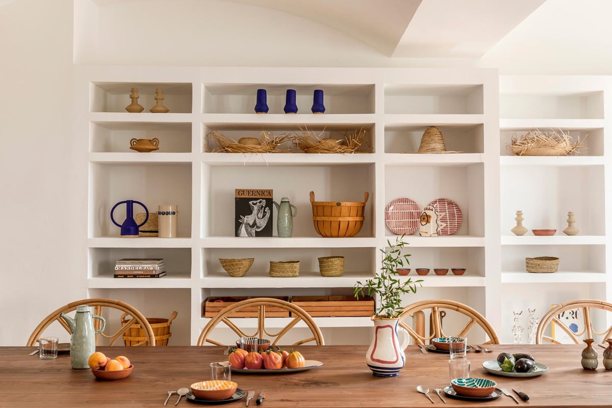 Menorca Experimental, Balearic Islands — Dining room with white built-in shelving, wooden table, woven chairs, and curated objects in natural materials.
