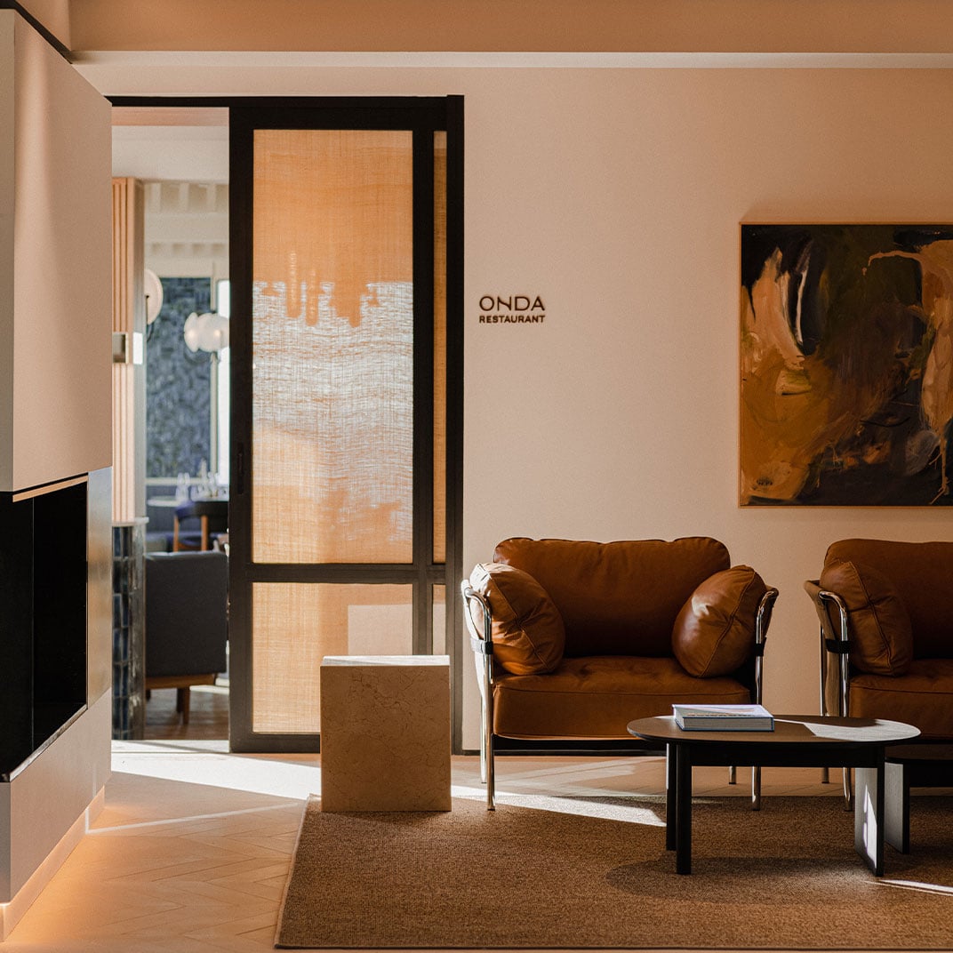 Aethos Ericeira, Portugal — Minimalist lounge with cognac leather armchairs, terracotta walls, black-framed glass door, and warm natural light filtering through textured screens.