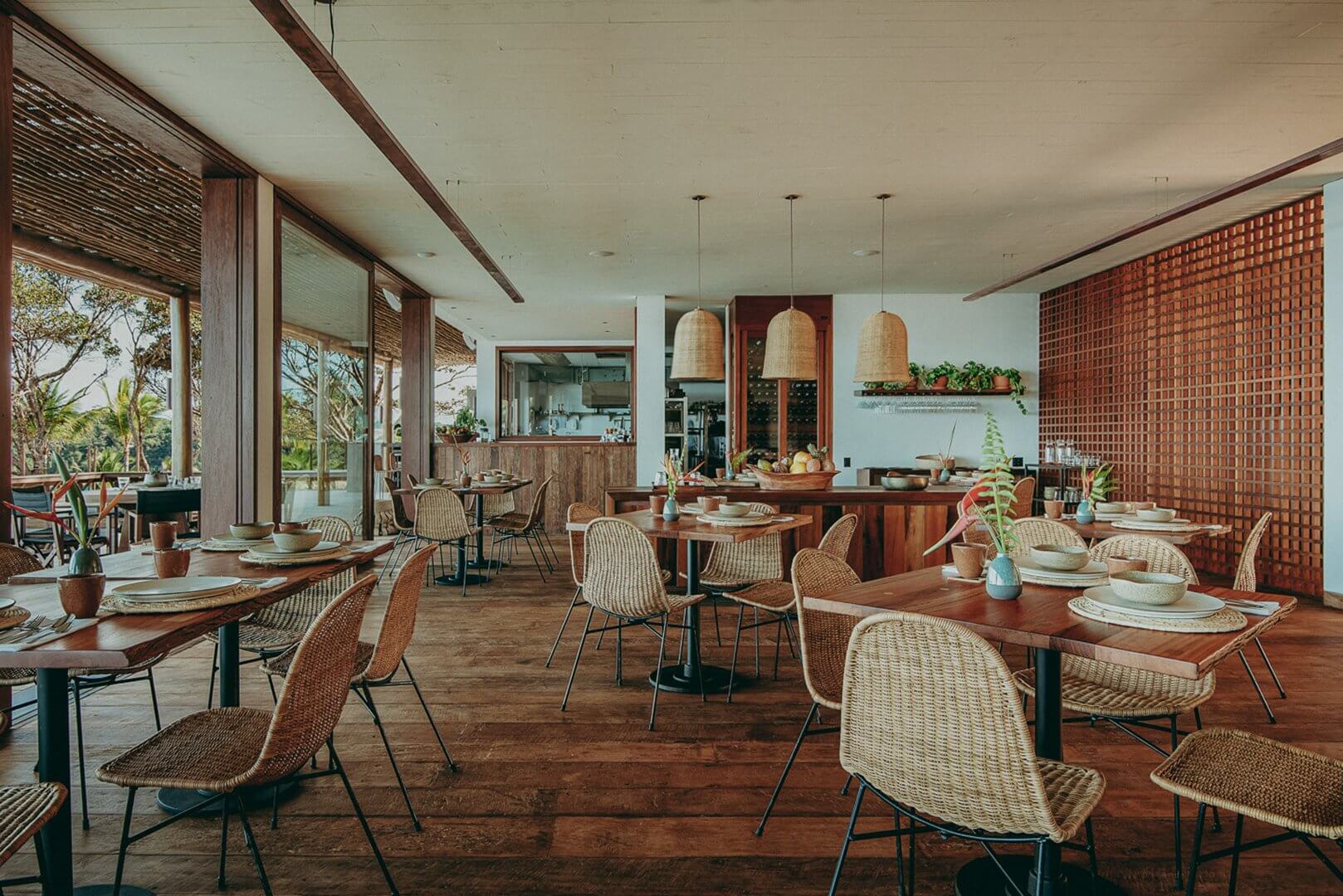 Barracuda Restaurant, South America — Woven rattan chairs, timber tables, terracotta brick screen wall, pendant lights, open kitchen, tropical garden views, warm minimalist interior.
