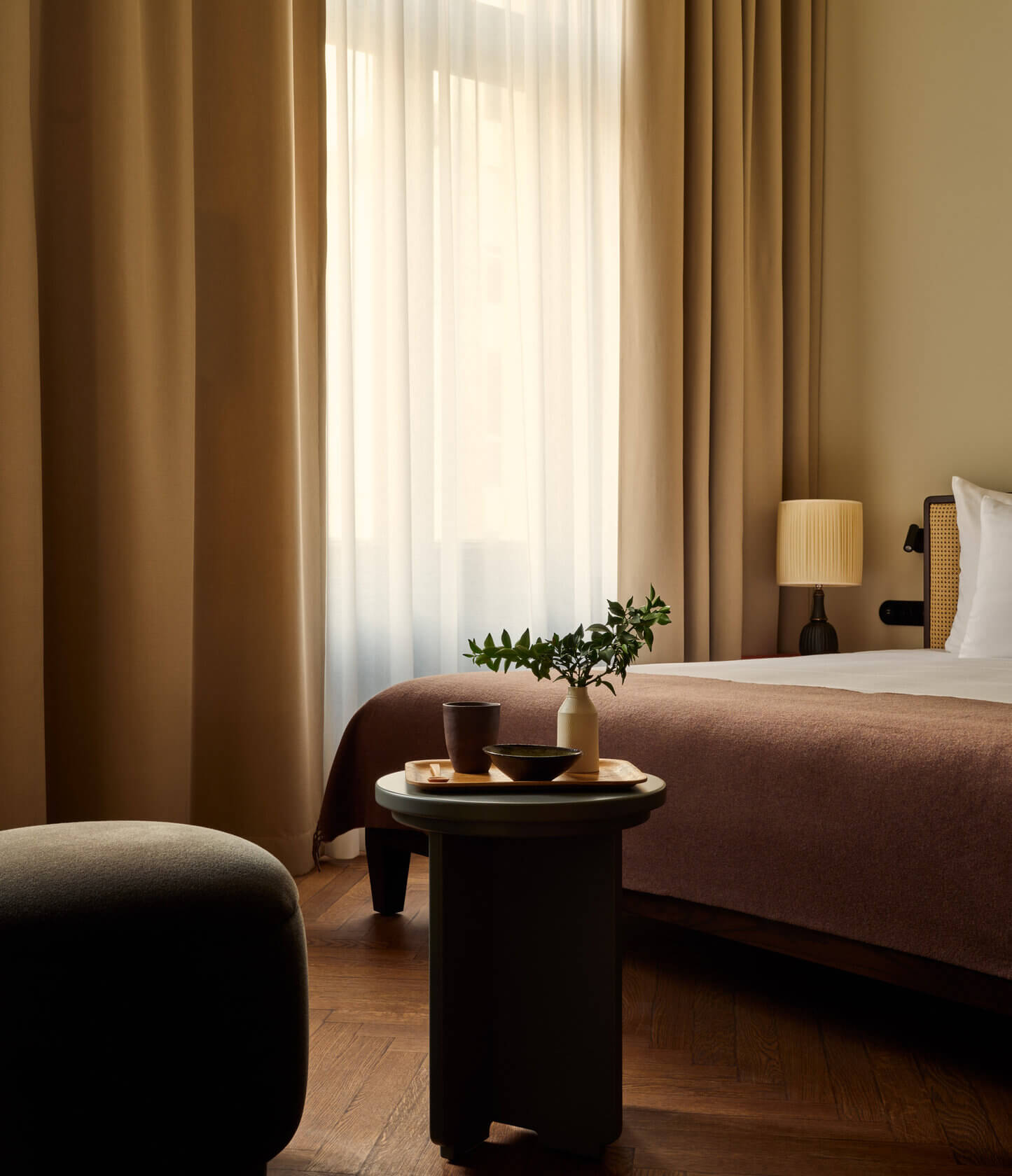 Château Royal, Europe — Minimalist hotel bedroom with warm earth tones, natural light through sheer curtains, dark side table with fresh greenery, and refined furnishings in neutral palette.