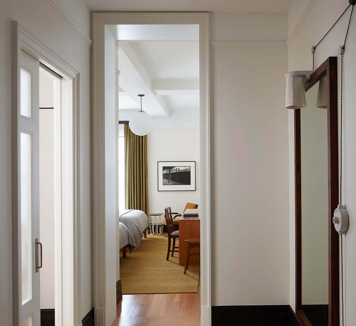 Nine Orchard, North America — Hallway with cream walls, dark baseboards, and warm wood flooring opening to a mid-century modern bedroom with olive curtains and natural light.