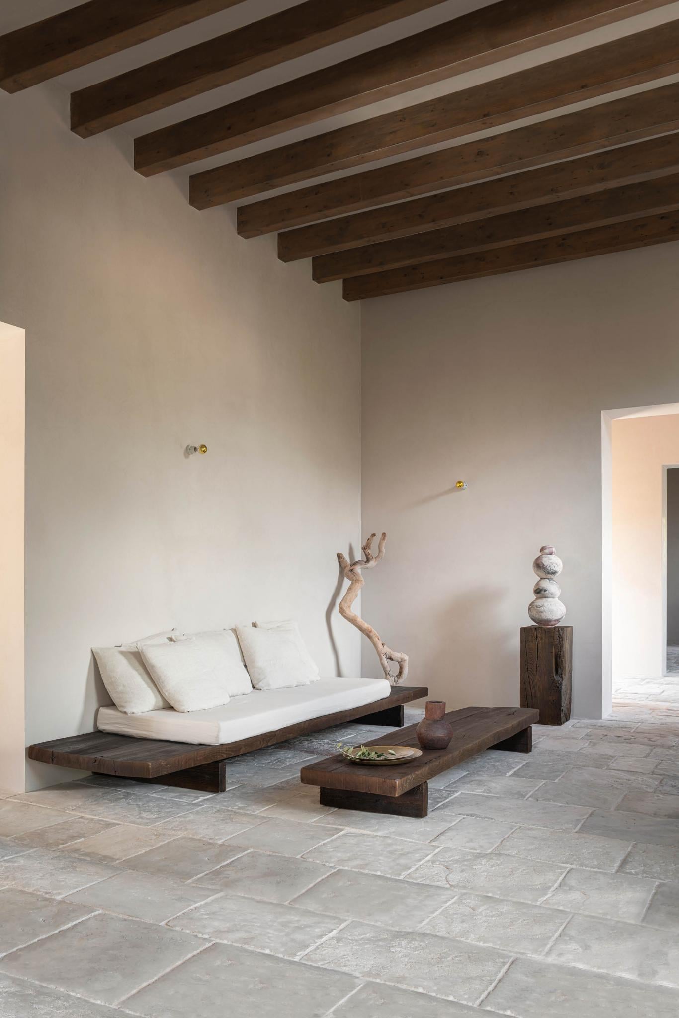 Can Gaspar, Balearic Islands — Minimalist living room with cream sofa, dark wood beams, stone flooring, driftwood sculpture, and sculptural stone stack on wooden plinth.