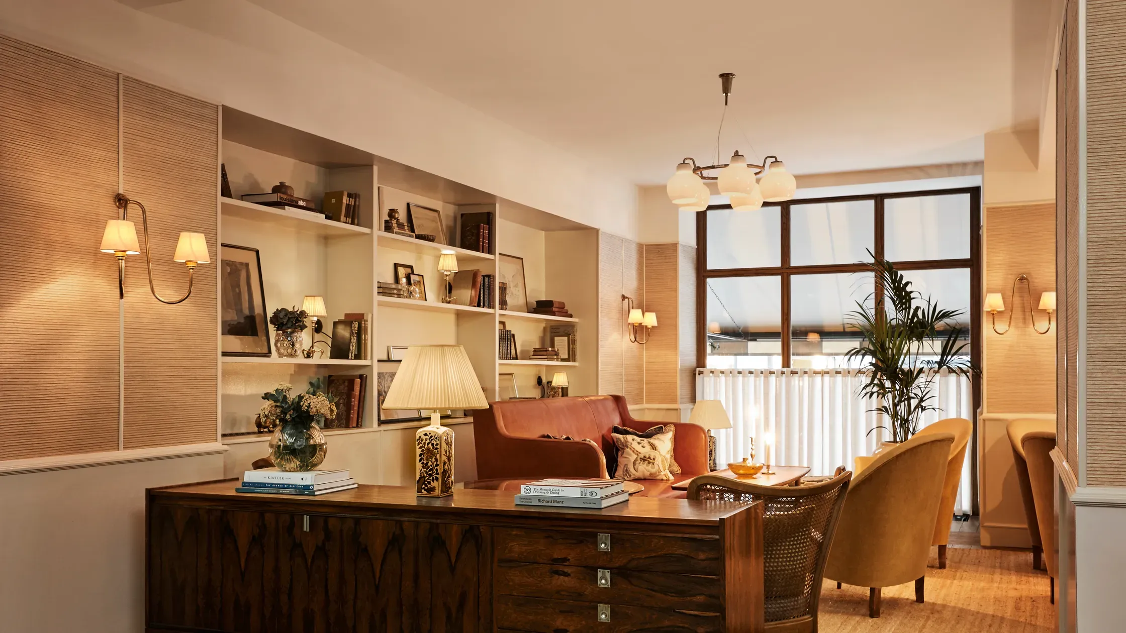 Hotel Sanders, Copenhagen, Denmark — Warm-lit executive suite with walnut desk, ochre velvet seating, built-in shelving, and floor-to-ceiling windows overlooking waterfront.