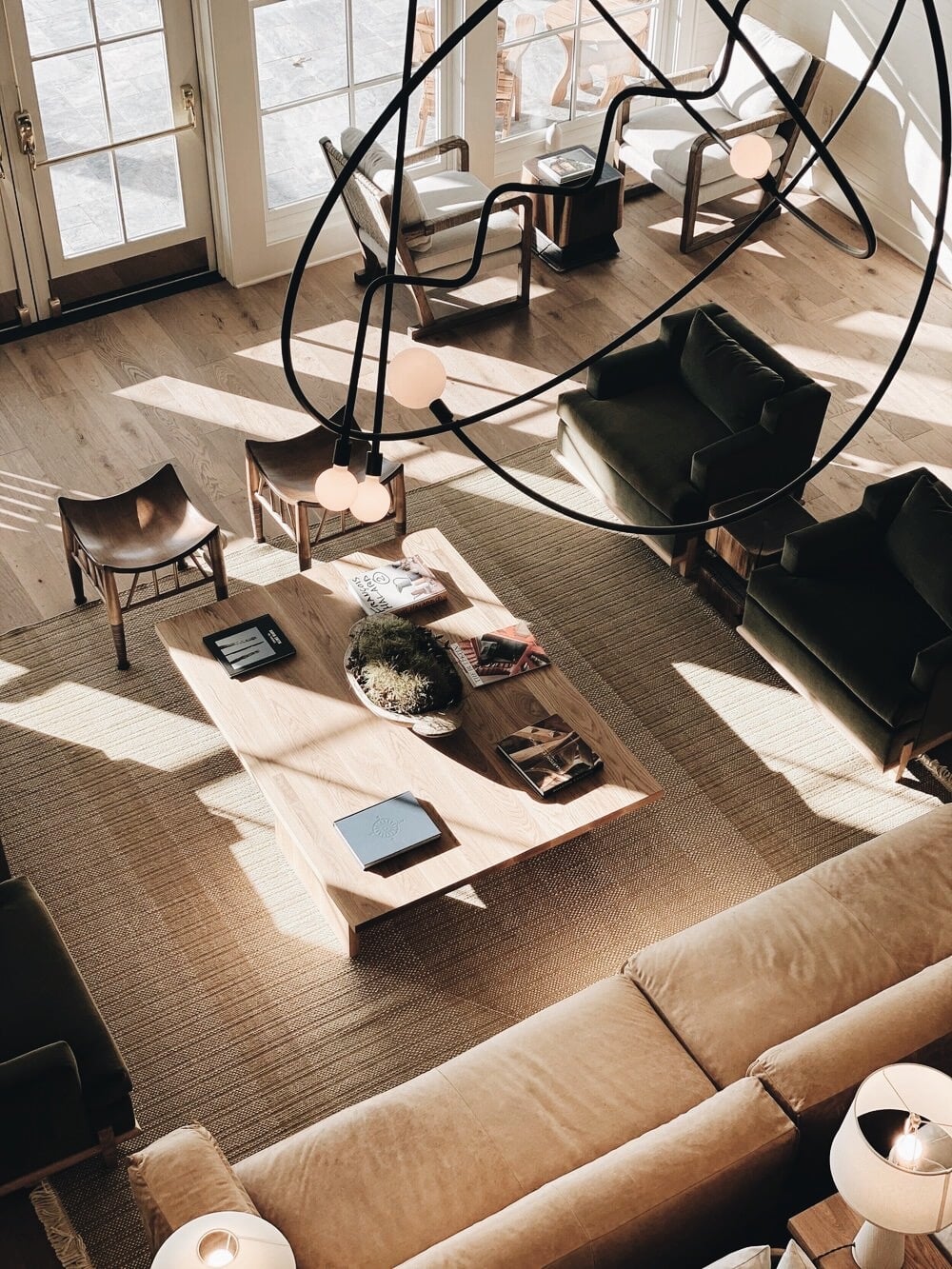 The Lake House, North America — Modern living room with floor-to-ceiling windows, sculptural black pendant lights, natural wood coffee table, neutral furnishings, and expansive views flooded with daylight.