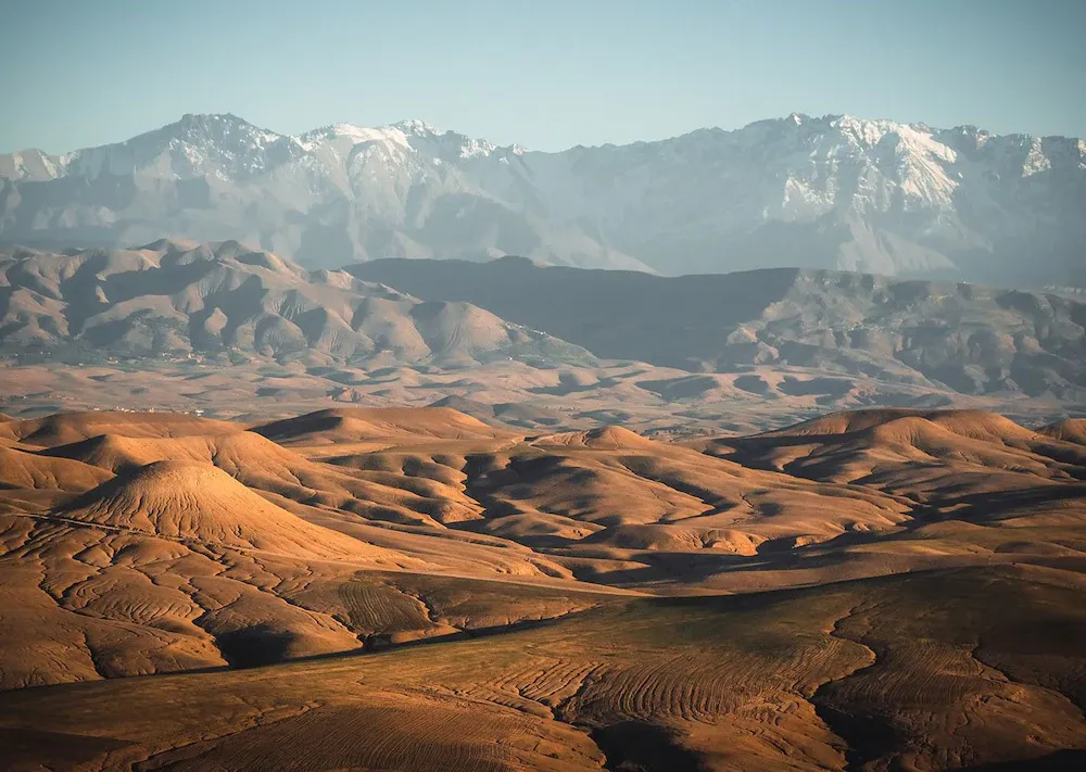 Scarabeo Camp, Africa — Rolling desert hills with terraced agricultural patterns, snow-capped mountains beyond, warm golden light casting deep shadows across arid landscape.