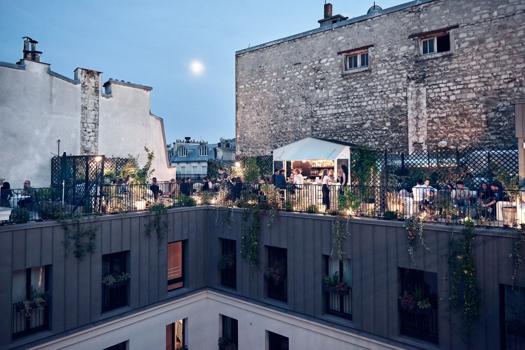 The Shed, Europe — Rooftop terrace with industrial stone buildings, candlelit bar tent, guests mingling at dusk under moonlight, black metal railings with climbing ivy, modern courtyard hospitality venue.