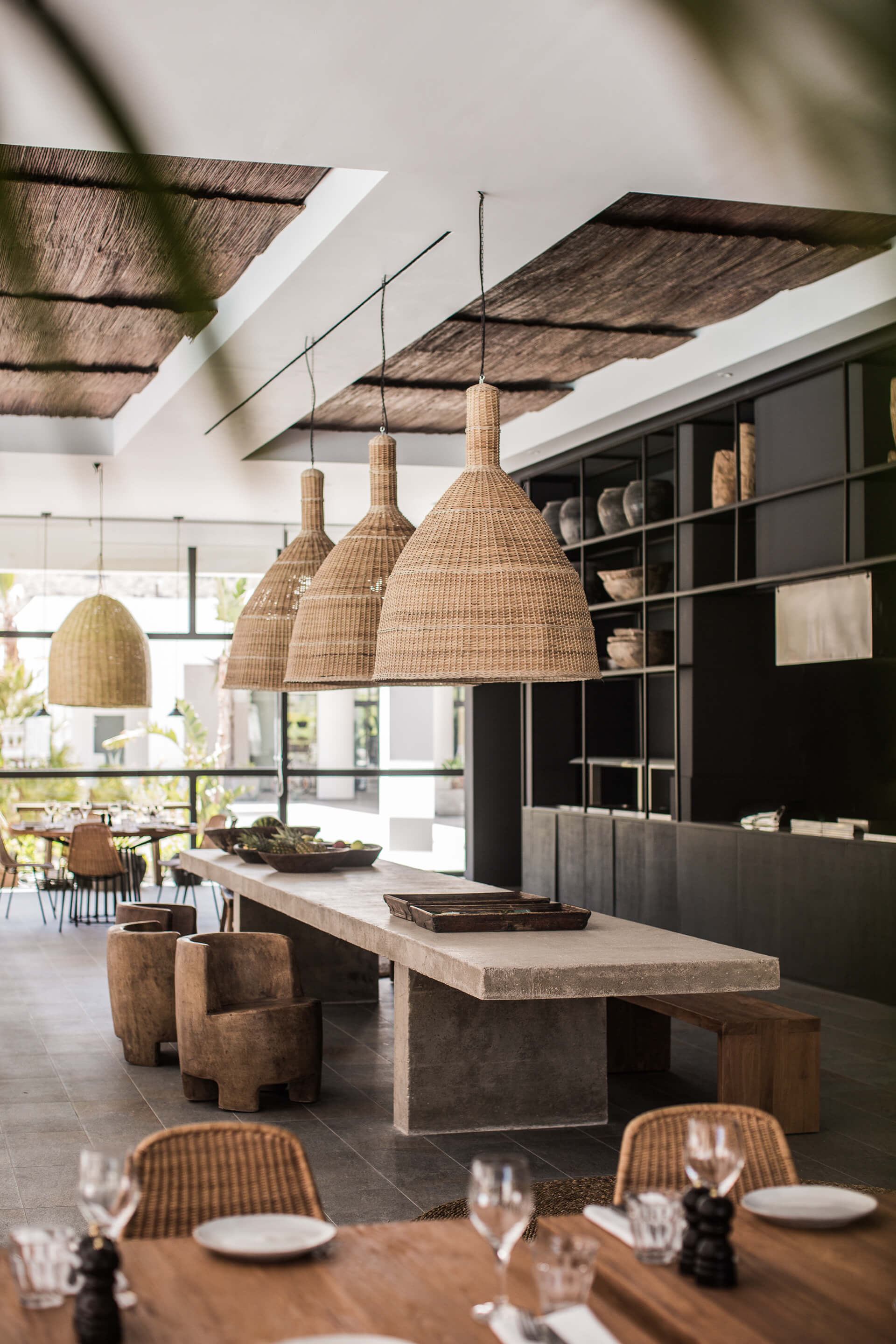 Casa Cook Rhodes, Europe — Natural woven pendant lights above concrete island counter, rattan chairs, black cabinetry, skylight with woven screens, modern Mediterranean dining space.