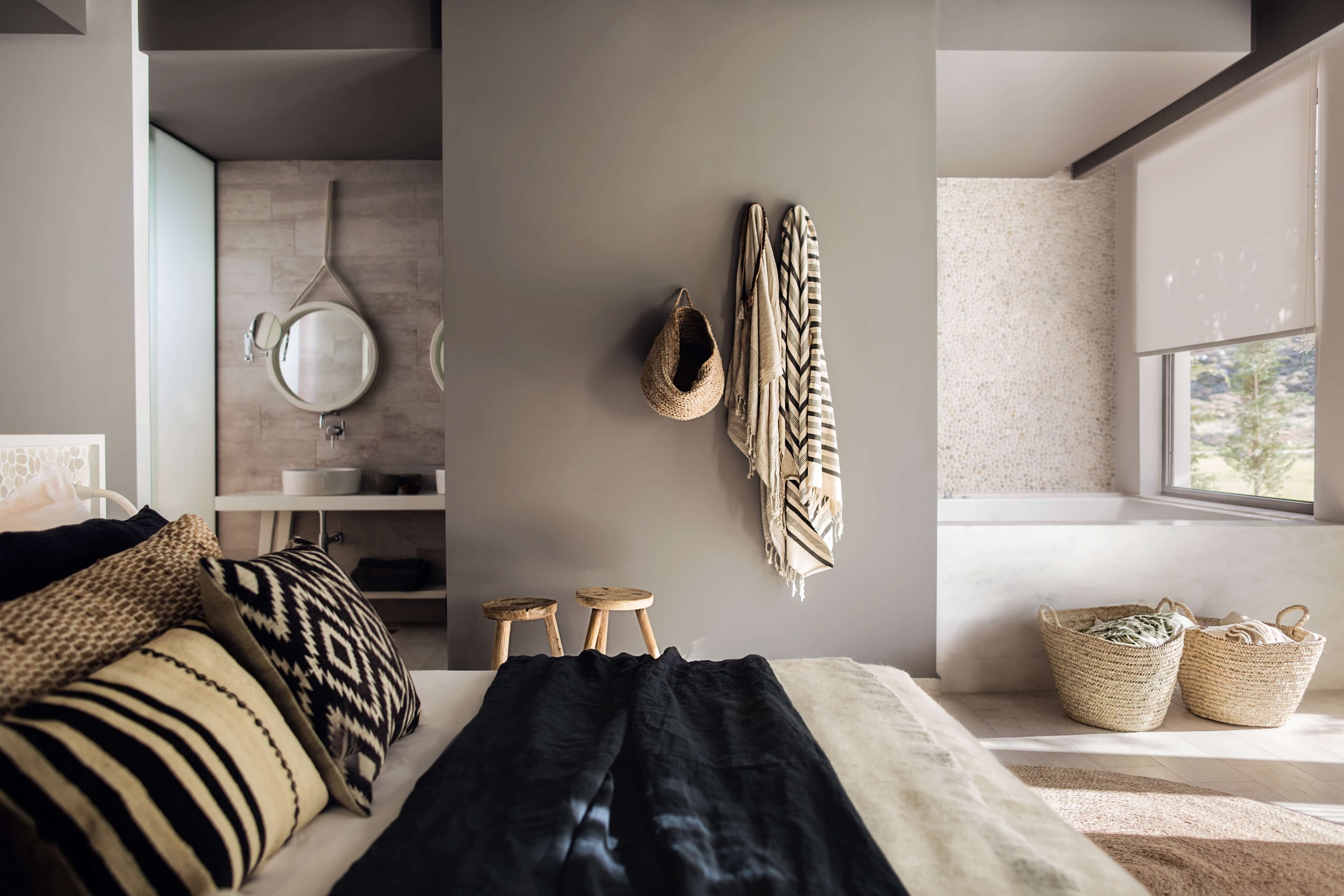 Casa Cook Rhodes, Europe — Minimalist bedroom with geometric patterned pillows, natural wood stools, woven baskets, and ensuite bathroom with circular mirror and neutral stone finishes.