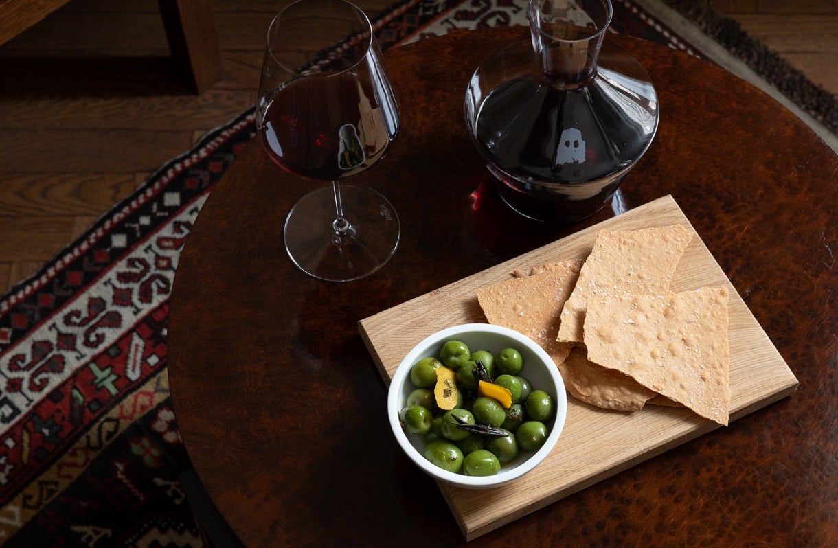 Château Royal, Europe — Wooden serving board with marinated green olives, crackers, and dark wine on mahogany table with ornate rug.