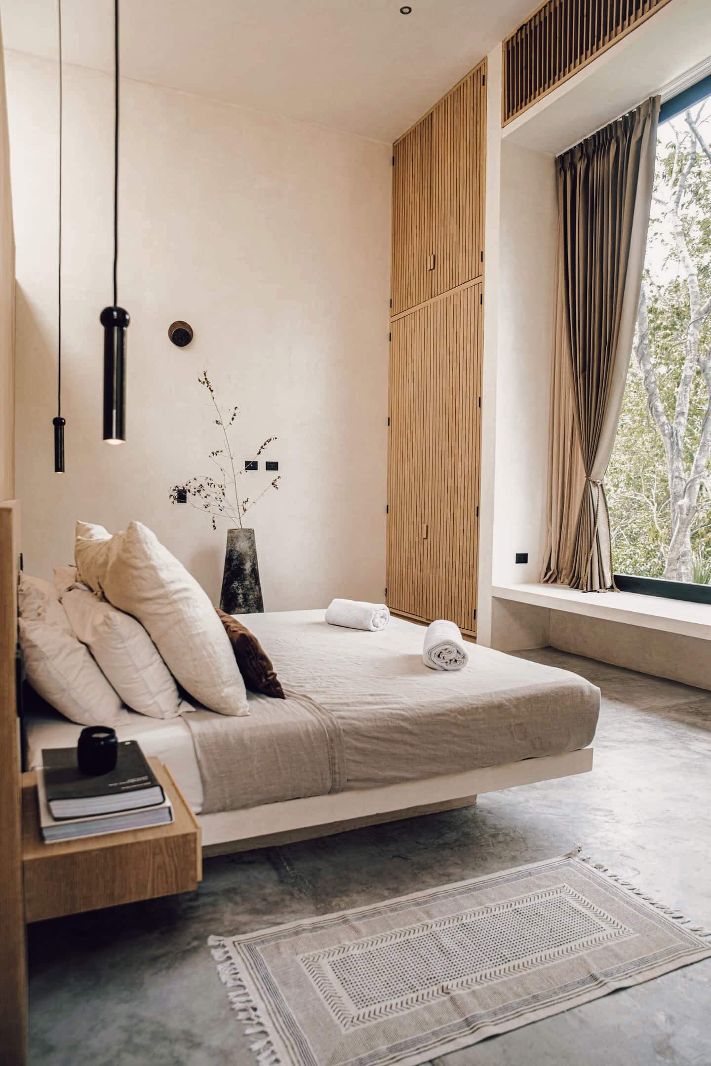 Villa Cava, Mexico — Minimalist bedroom with natural linen bedding, wooden slat wardrobe, pendant lights, and floor-to-ceiling windows overlooking forested landscape.