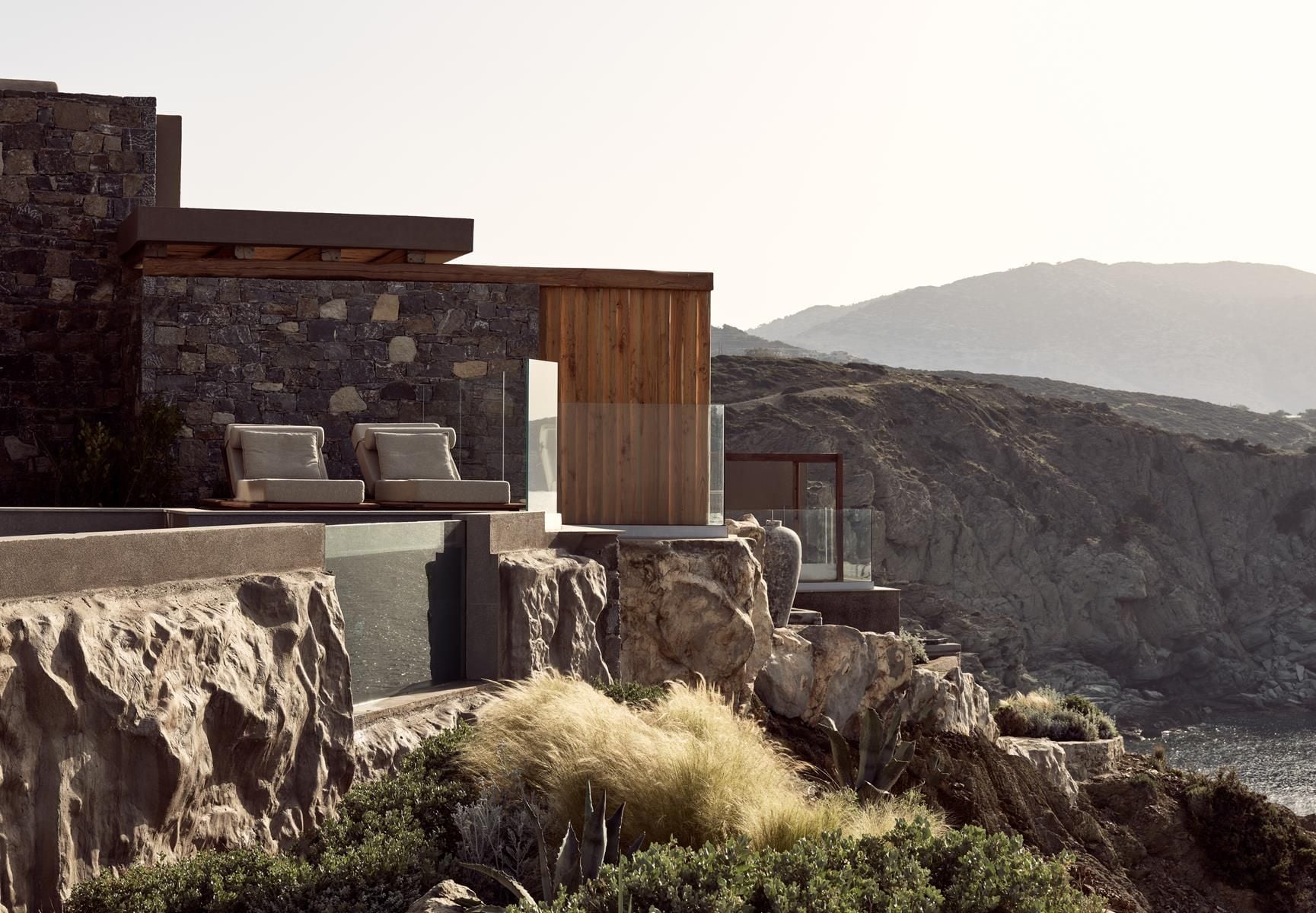 ACRO Suites, Europe — Modern stone and timber pavilion perched on desert rocks with lounge seating, mountains beyond, warm natural light.