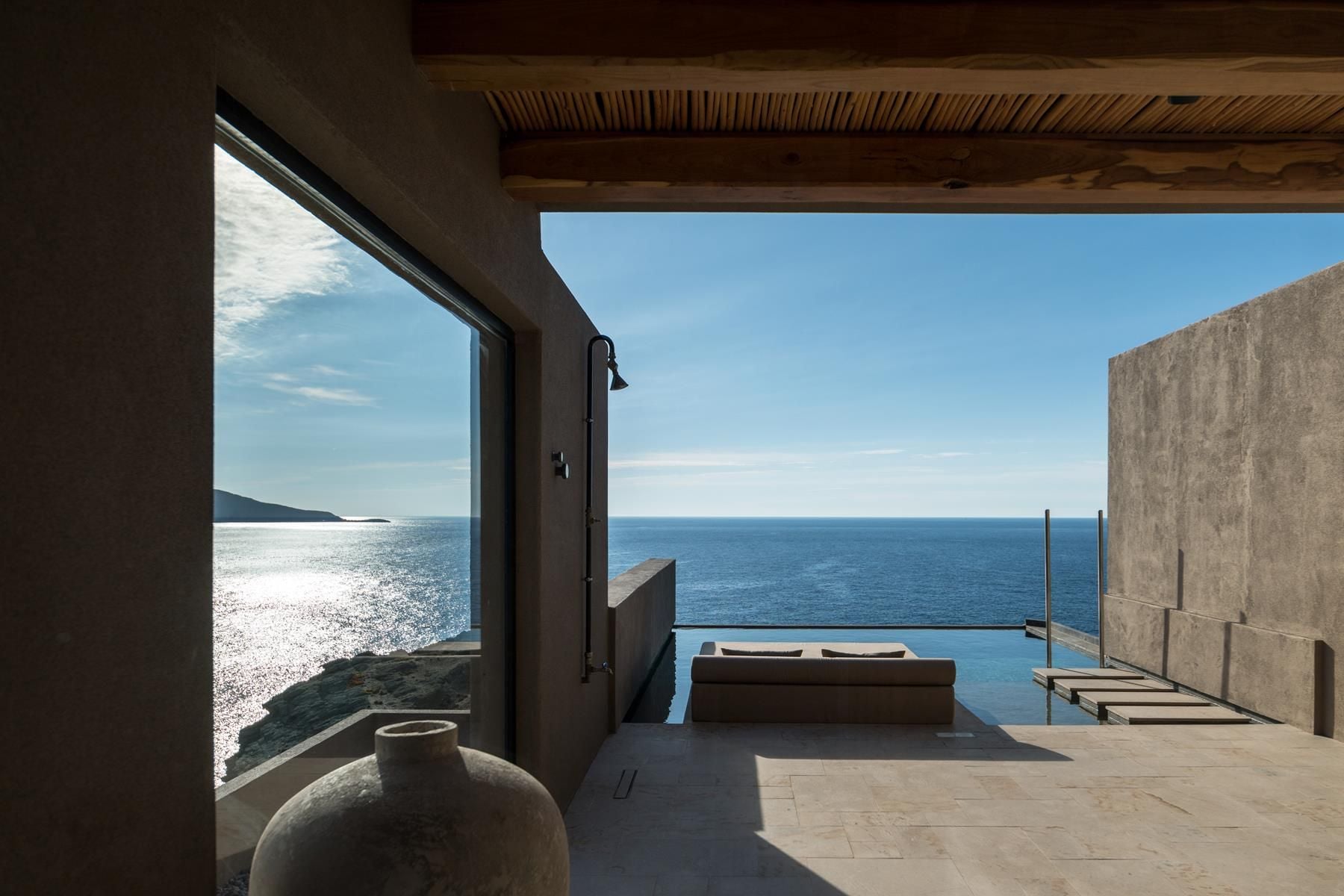 ACRO Suites, Europe — Seaside terrace with concrete overhang, minimalist bench seating, glass railing overlooking calm blue ocean and distant headland under clear sky.