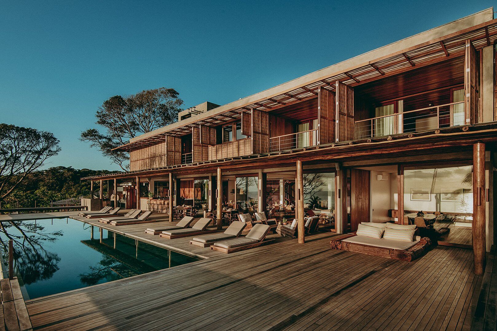 Barracuda Hotel & Villas, South America — Waterfront timber pavilion with lounge chairs, wooden deck, and floor-to-ceiling glass overlooking still water at dusk.