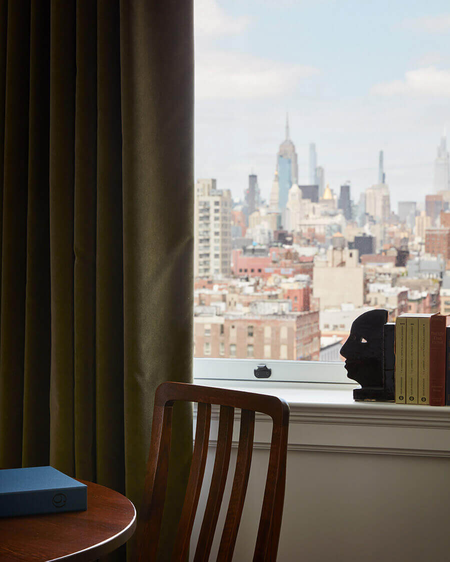 Nine Orchard, New York, United States — Midcentury chair and wooden desk with city view of Manhattan skyline framed by olive curtains.