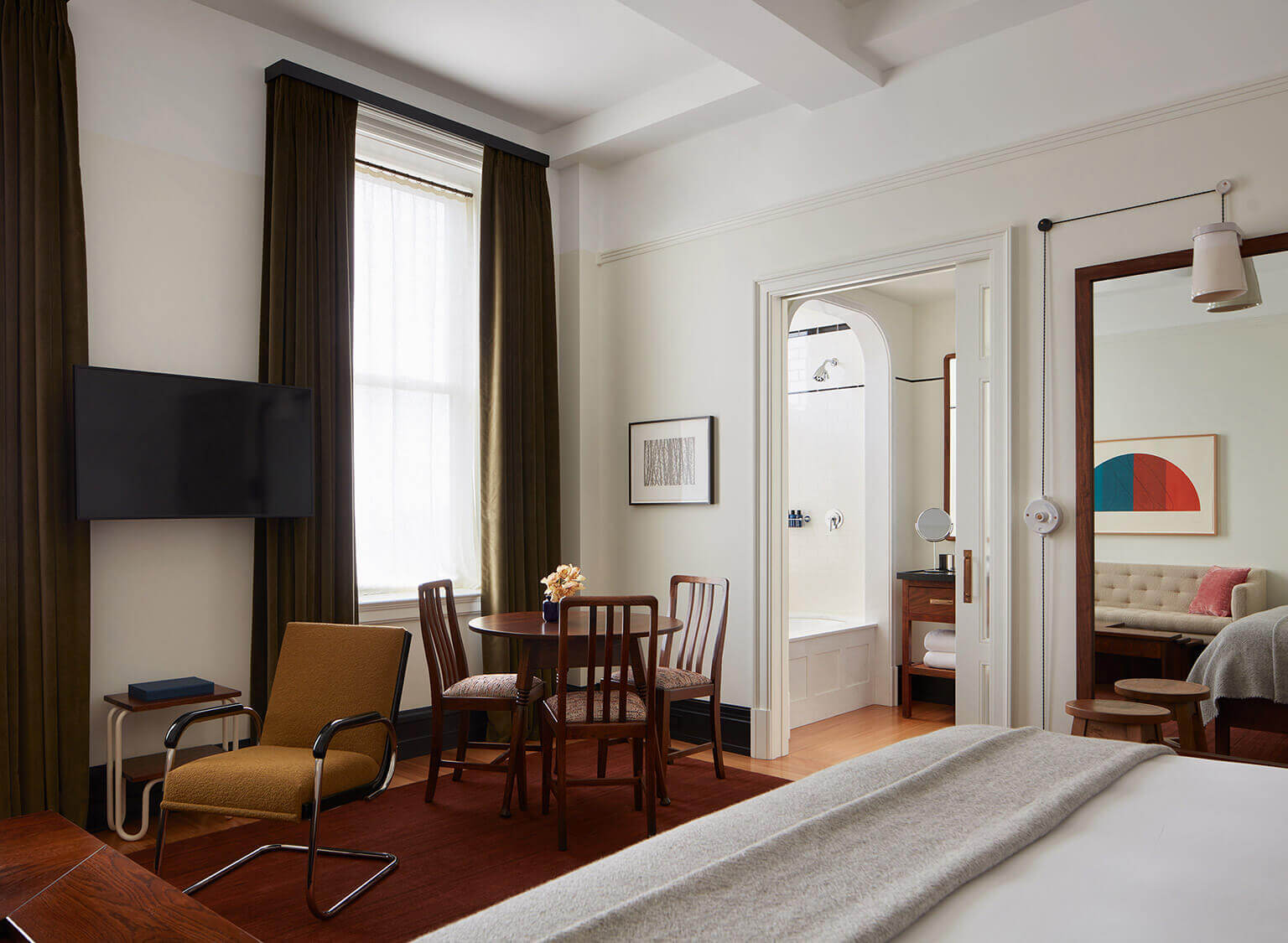 Nine Orchard, North America — Midcentury modern suite with wood furnishings, cognac leather chair, round dining table, and bathroom ensuite visible through open doorway.