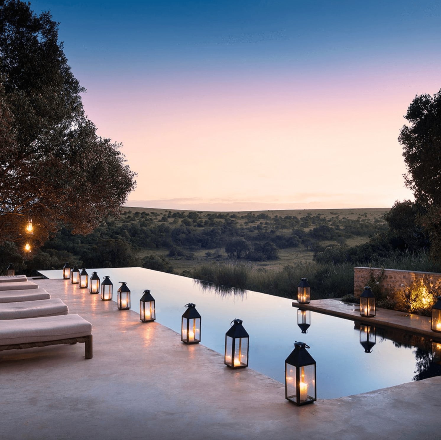 Arijiju Retreat, Africa — Infinity pool with candlelit lanterns overlooking savanna landscape at dusk, minimalist architecture blending stone and water.