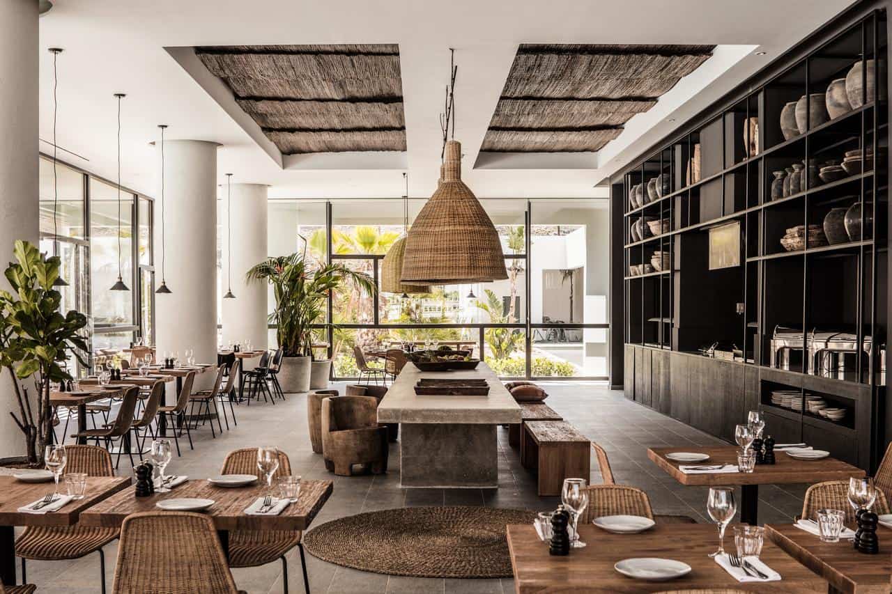 Casa Cook – Rhodes — Casa Cook Rhodes — Modern restaurant interior with woven ceiling panels, black shelving, natural wood tables, rattan chairs, and floor-to-ceiling windows overlooking gardens.