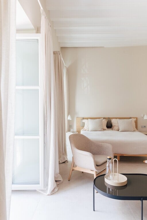 Plácido y Grata, Europe — Minimalist bedroom with natural light, wooden bed frame, neutral textiles, linen curtains, and modern furnishings in warm beige tones.