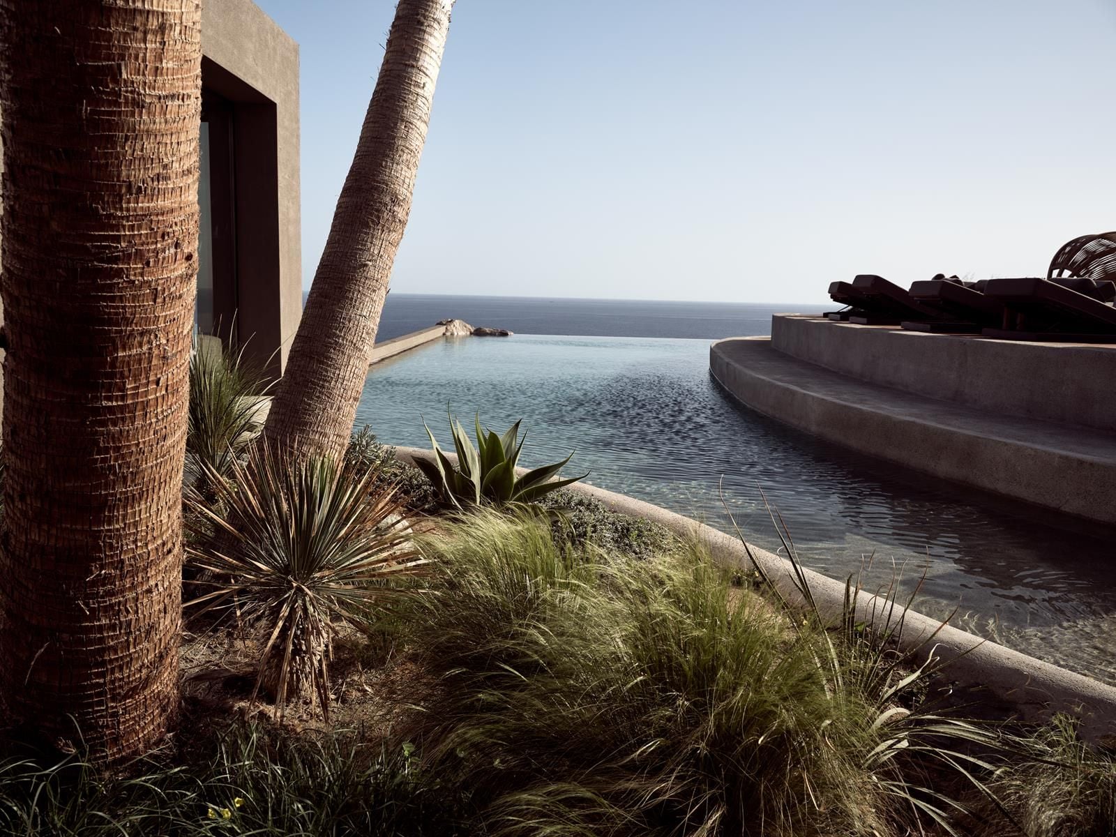 ACRO Suites, Europe — Infinity pool overlooking Mediterranean Sea, modernist concrete architecture with palm trees and desert landscaping, clear horizon light.