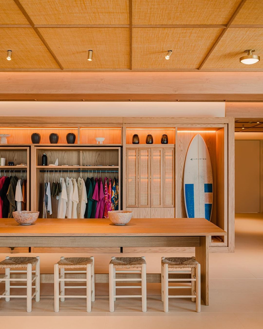 Aethos Ericeira, Portugal — Warm timber retail space with surfboard display, natural linen clothing racks, woven seating, and coffered wood ceiling with ambient lighting.