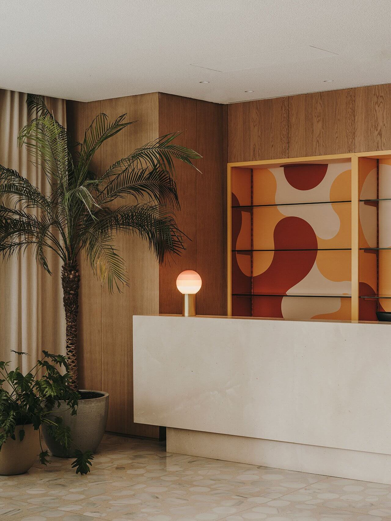 The Standard Ibiza, Balearic Islands — Warm wood-paneled reception with geometric abstract artwork, palm plant, and minimalist white desk under soft lighting.