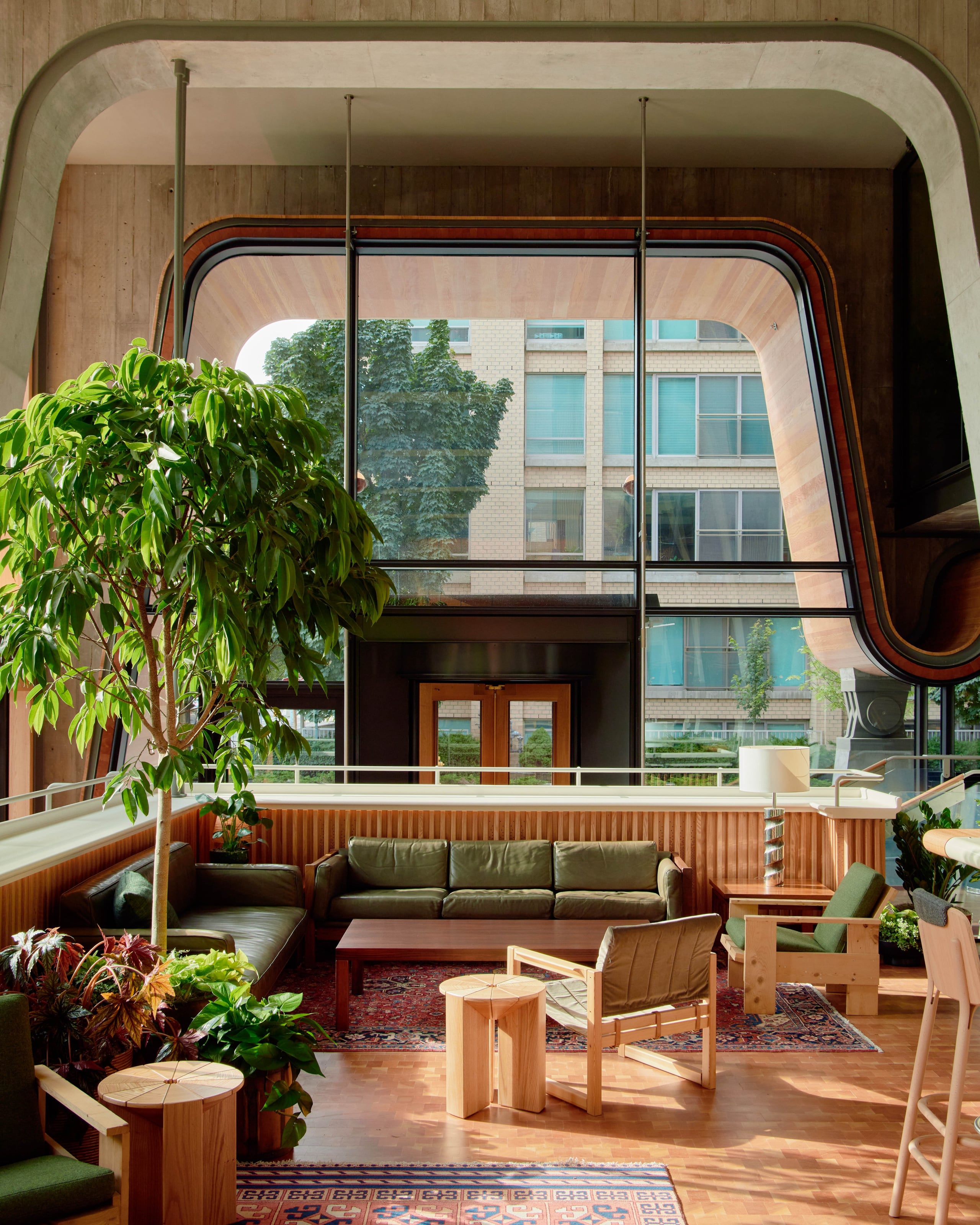 Ace Hotel Toronto, Canada — Mid-century modern lounge with soaring windows, green leather seating, wood furnishings, potted plants, and city views through sculptural concrete frame.