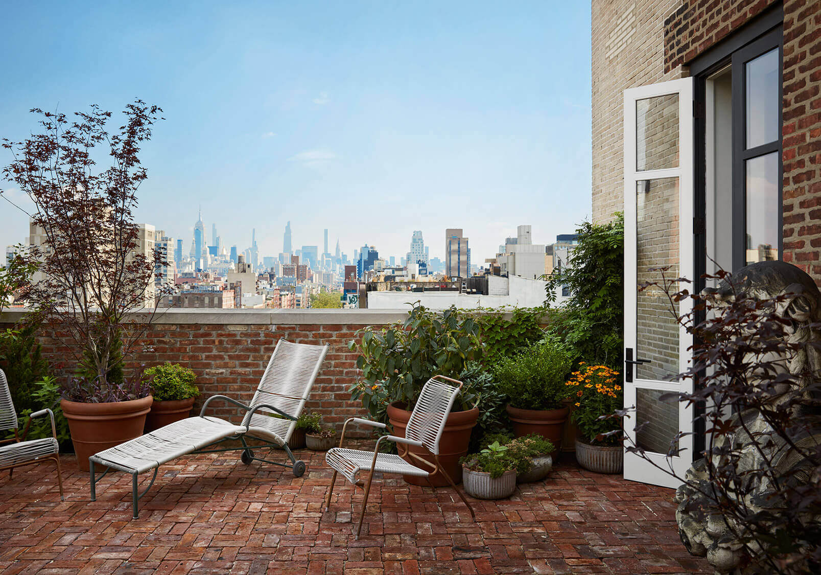 Nine Orchard, New York, USA — Rooftop terrace with lounge chairs, potted plants, and Manhattan skyline view across brick parapet.