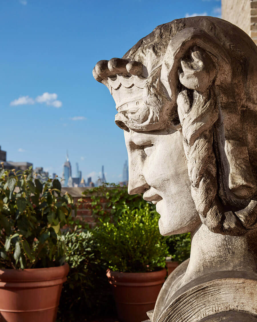 Nine Orchard, North America — Stone bust sculpture overlooking rooftop garden with Manhattan skyline, neoclassical architectural detail against clear blue sky.