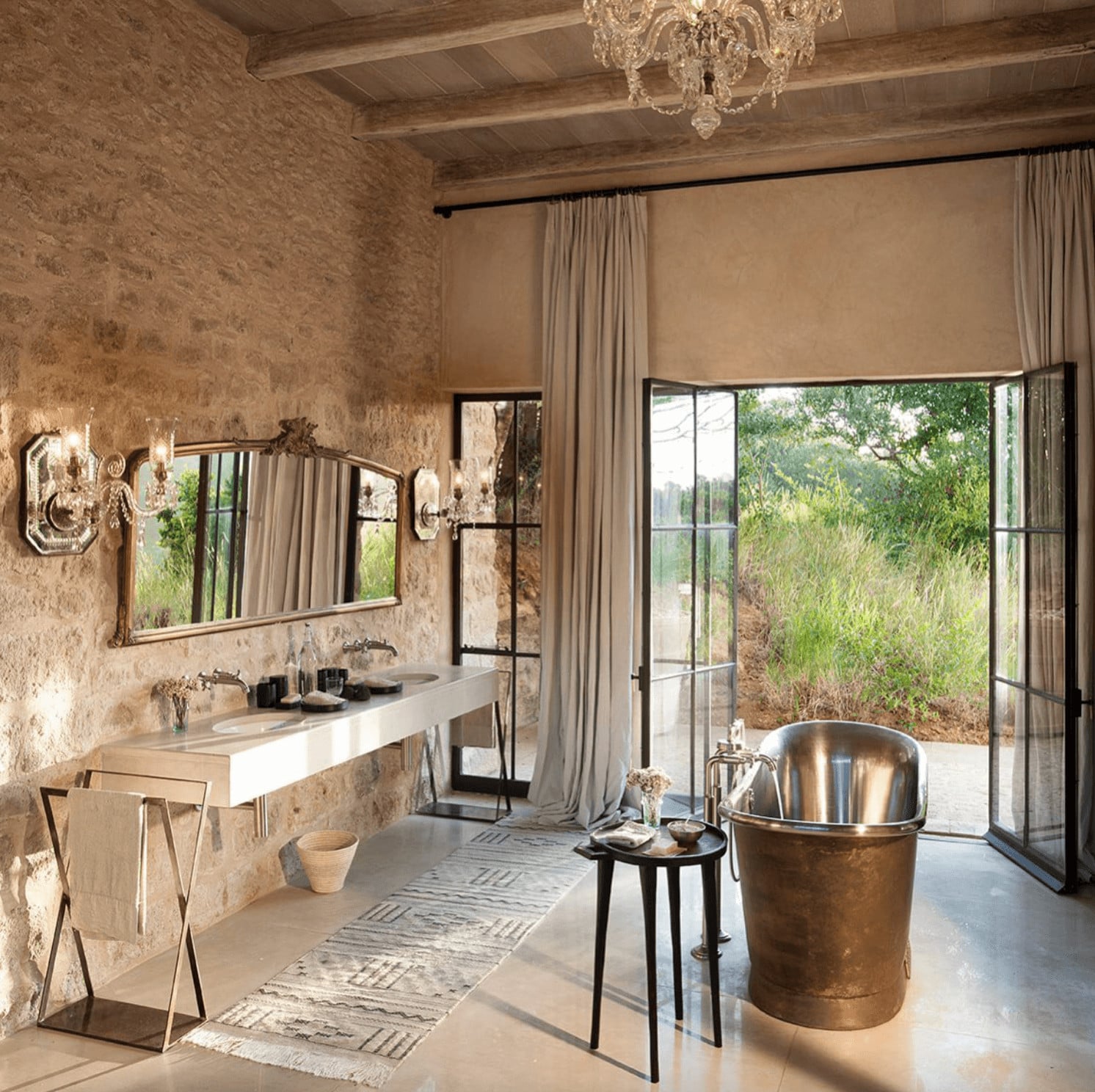 Arijiju Retreat, Africa — Spa bathroom with travertine walls, brass soaking tub, floating vanity, crystal chandelier, and garden views through black-framed doors.