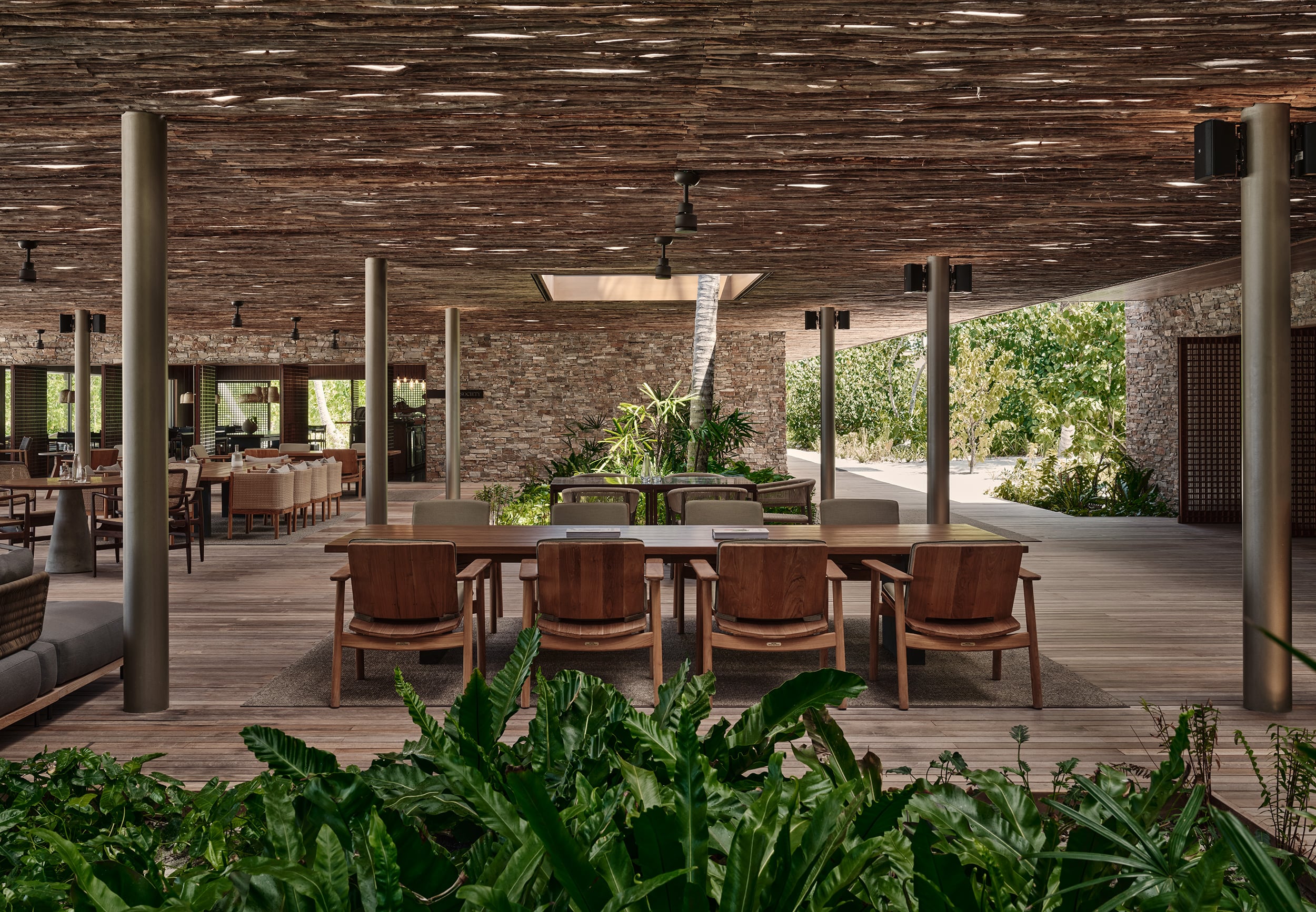 Patina Maldives, Asia — Open-air dining pavilion with timber deck, brick walls, concrete columns, and mid-century wooden chairs surrounding long table under textured ceiling.
