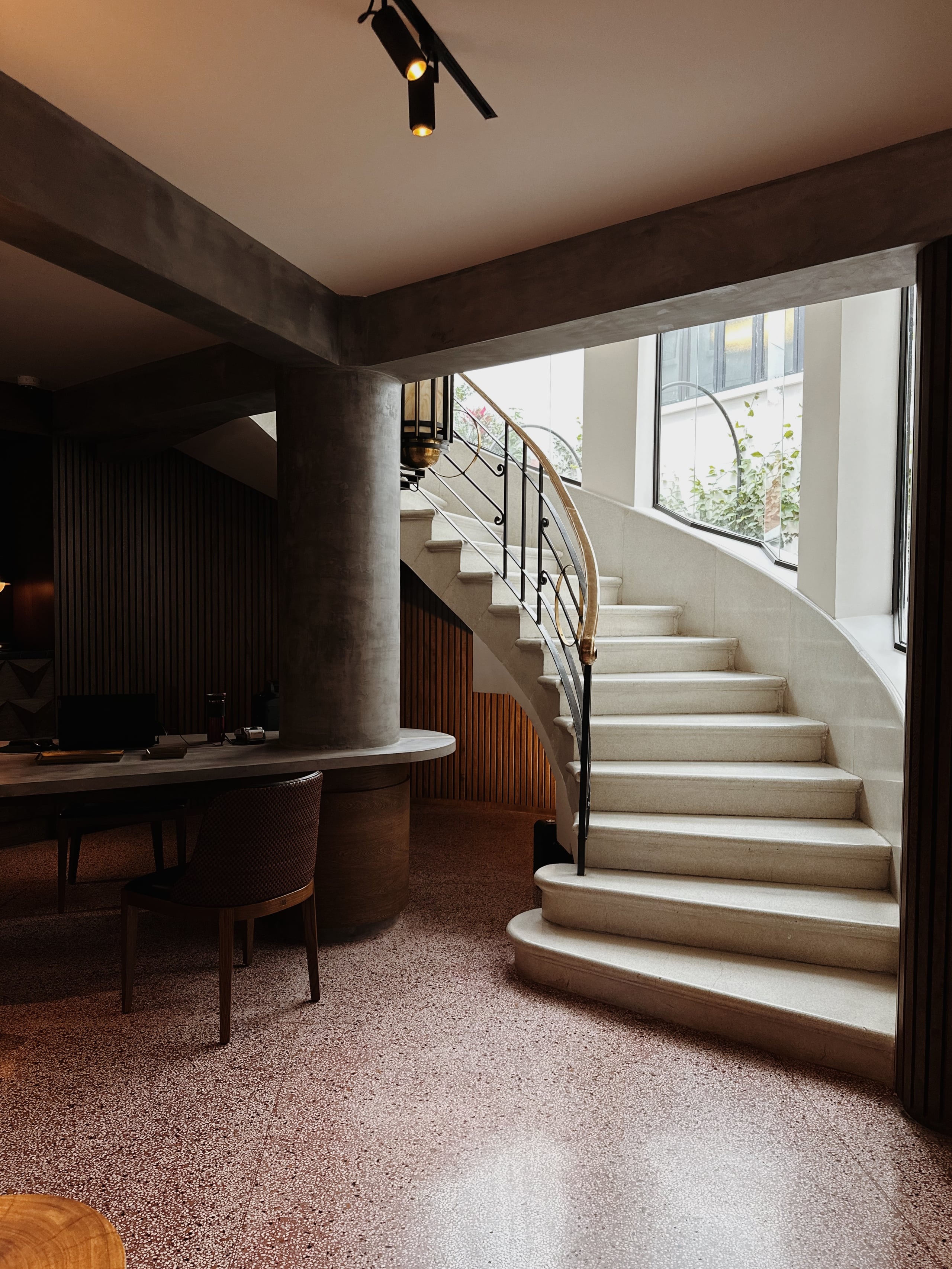 Campos Polanco, Mexico — Curved staircase with metal railing ascending past floor-to-ceiling windows, concrete column, terrazzo flooring, modern luxury interiors.
