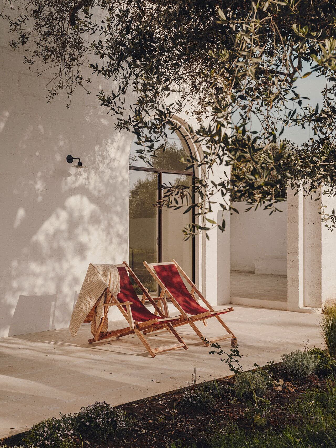 Borgo Gallana, Europe — Wooden deck chairs with red fabric beneath olive tree branches, casting shadows on limestone terrace adjoining modernist stone pavilion.