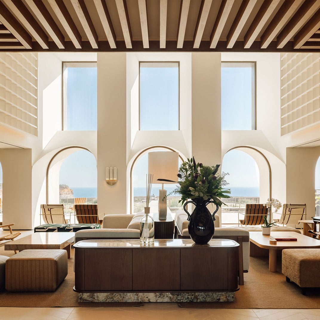 Aethos Ericeira, Portugal — Minimalist coastal lounge with arched alcoves, striped wooden ceiling, ocean views, and warm neutral palette with dark furnishings.