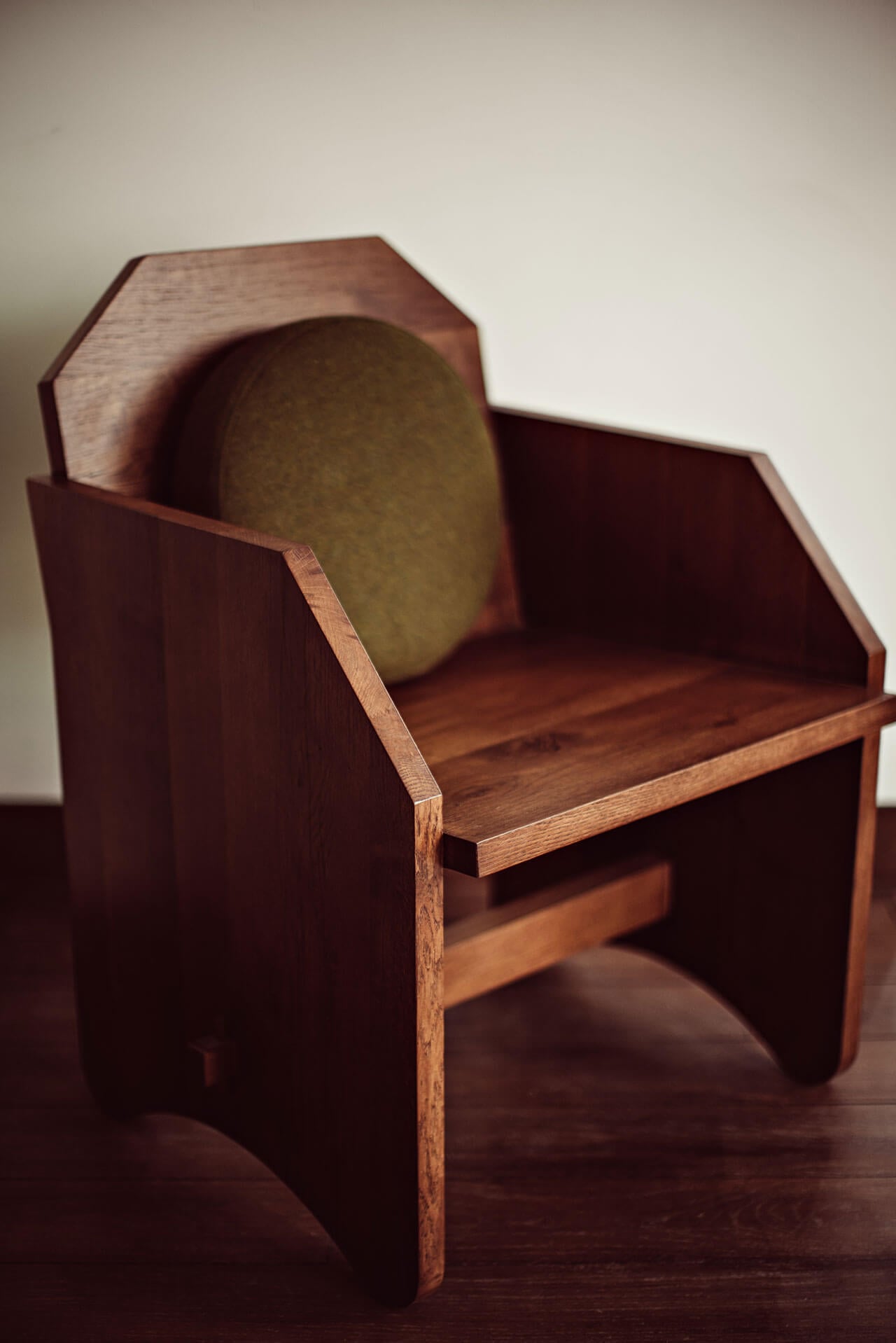 Académies et des Arts, Europe — Modernist wooden armchair with olive cushion, geometric form and warm wood grain detail against neutral backdrop.