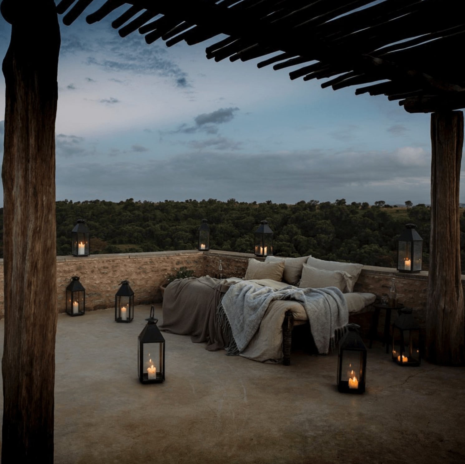 Arijiju Retreat, Africa — Elevated outdoor bedroom terrace with wooden pergola, stone walls, neutral linens, candlelit lanterns overlooking savanna landscape at dusk.