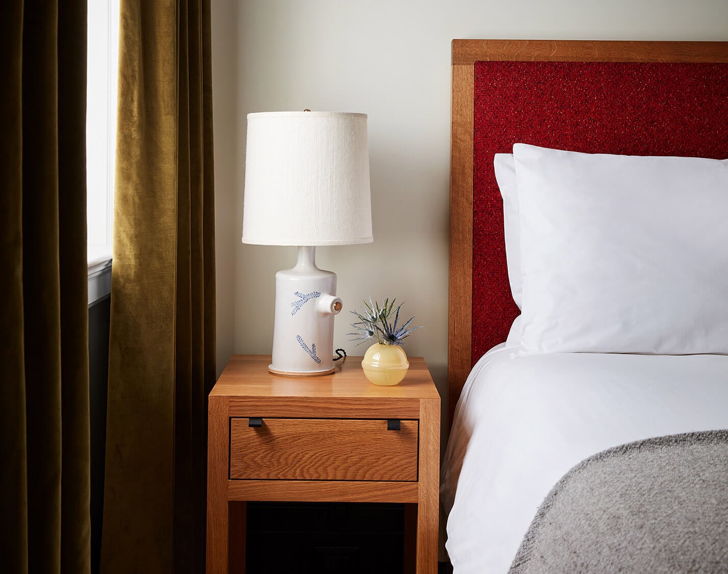 Nine Orchard, North America — Bedside table with ceramic lamp, yellow vessel, and dried botanicals beside bed with burgundy headboard and white linens.