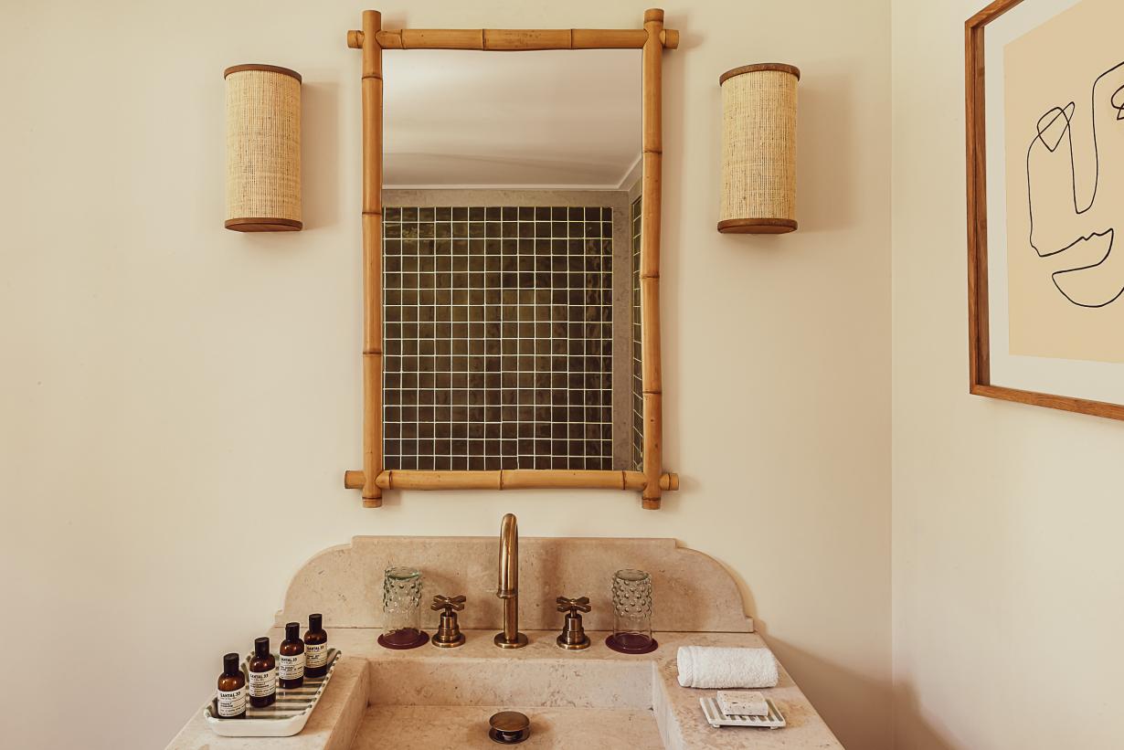 Hôtel Lou Pinet, Europe — Travertine bathroom sink with brass fixtures, bamboo-framed mirror, woven wall sconces, and natural light highlighting minimalist luxury design.