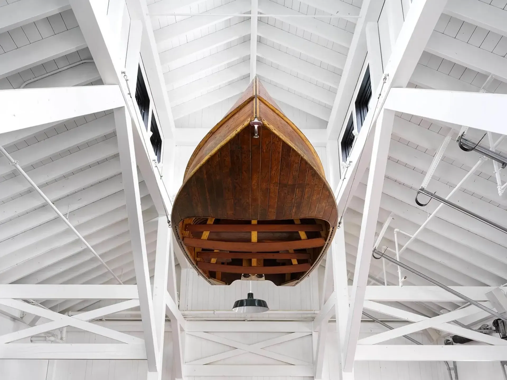 The Lake House, North America — Wooden rowing boat suspended in vaulted white timber boathouse interior with exposed beam ceiling and nautical hardware.