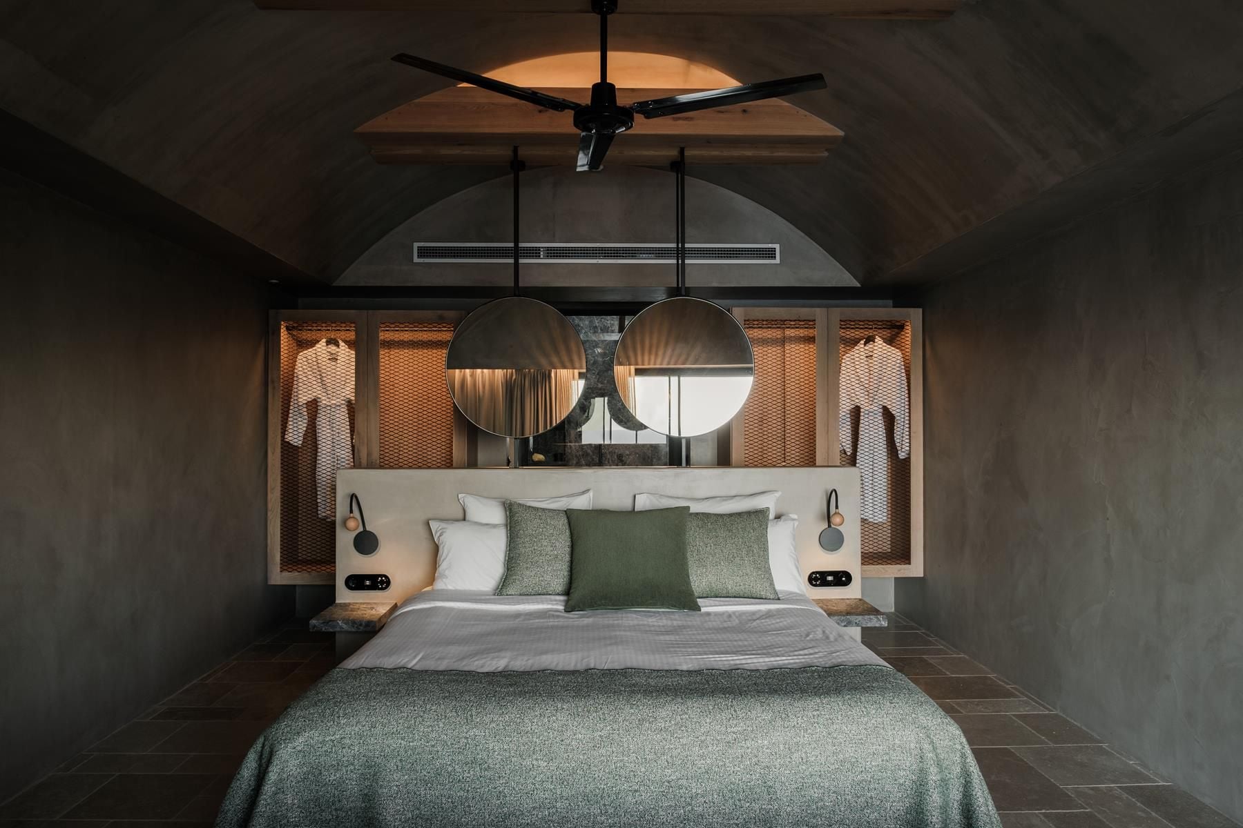 ACRO Suites, Europe — Modern hotel bedroom with arched ceiling, concrete walls, round mirrors, pendant lights, green textiles, and ceiling fan with warm ambient lighting.