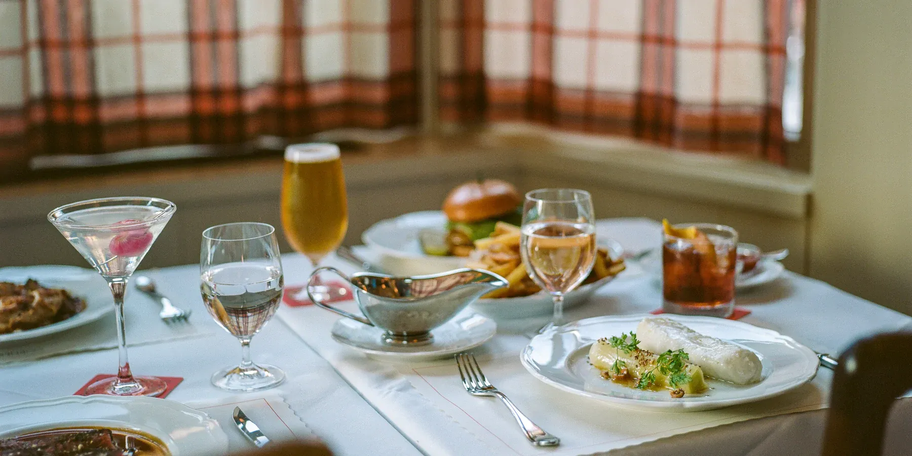 Corner Bar, North America — Elegant fine dining table setup with plated fish entrée, cocktails, and golden beer under natural light from tall windows.