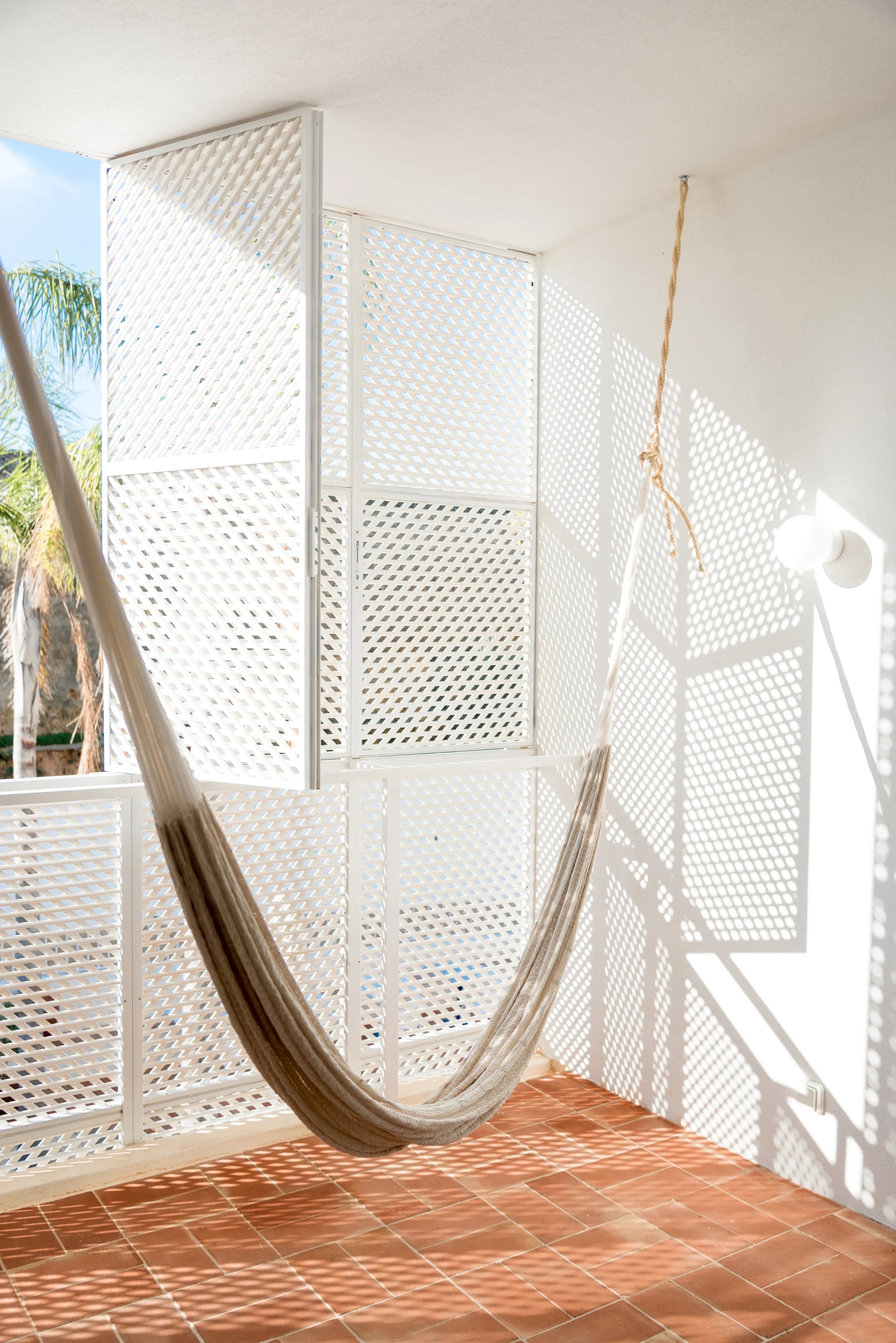 Casa Mãe, Europe — Minimalist outdoor room with white perforated metal screens, terracotta floor, and draped hammock casting geometric shadows in bright sunlight.